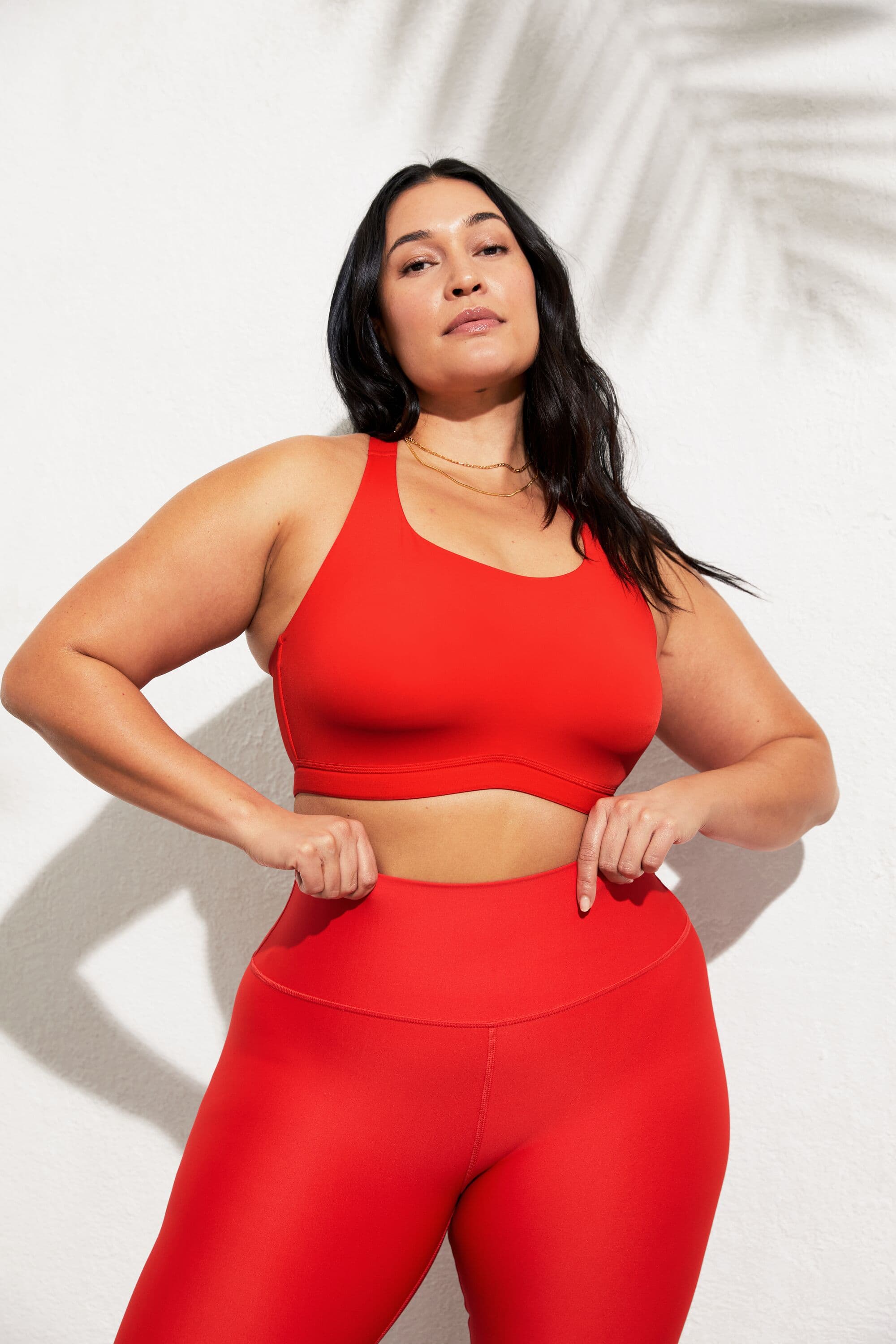 A woman wearing a high impact bra with a pair of red high-waist leggings while posing with her hands at her hips in front of a white wall with a palm frond shadow in the background.  