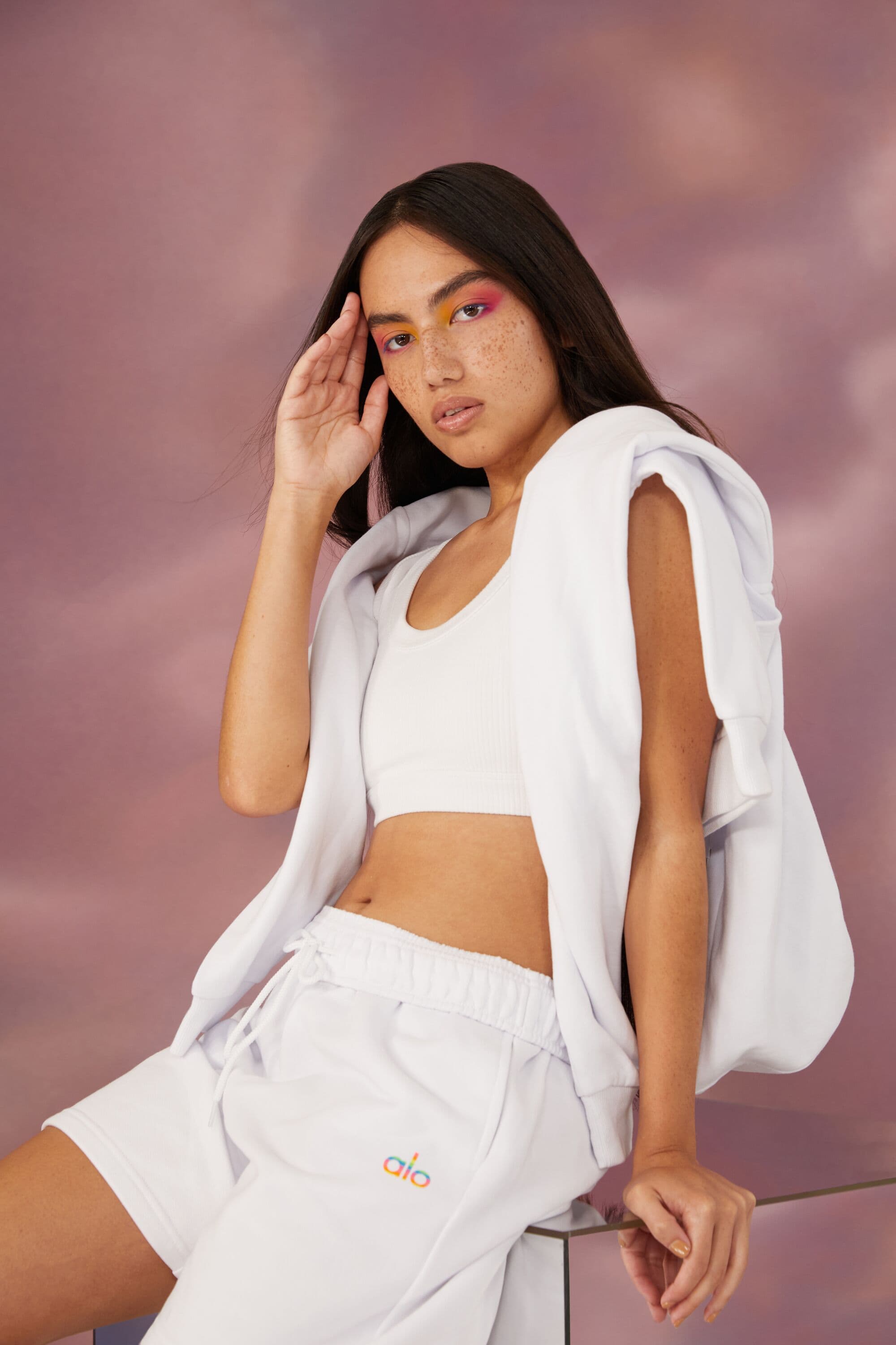 A woman wearing an all-white outfit from Alo’s 2022 Pride Collection while posing on a mirrored block in front of a pink cloud backdrop. 