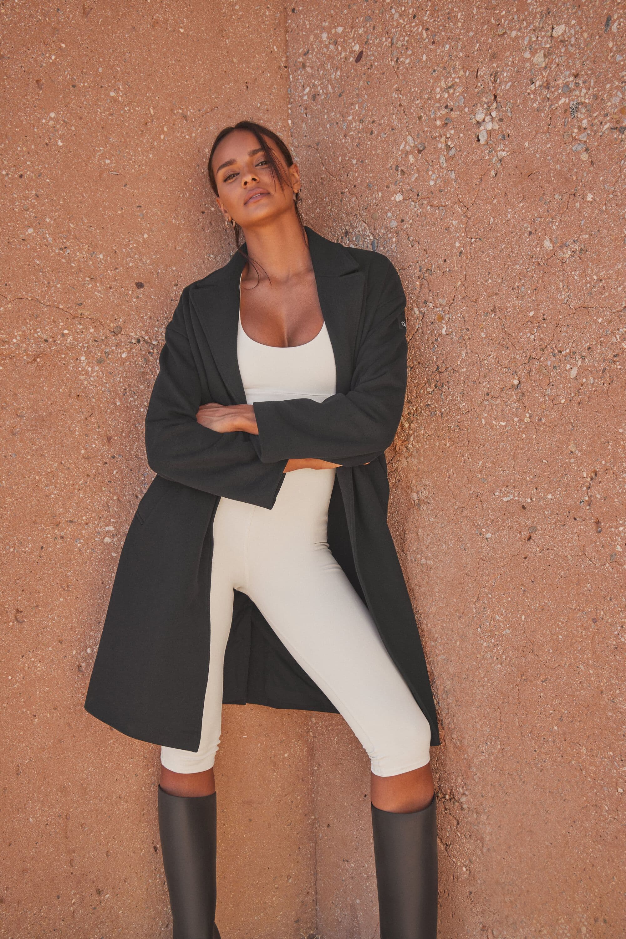 A woman wearing an ivory capri length activewear onesie with a full-length trench coat and knee-high boots posing cross-armed in front of a clay-colored wall. 