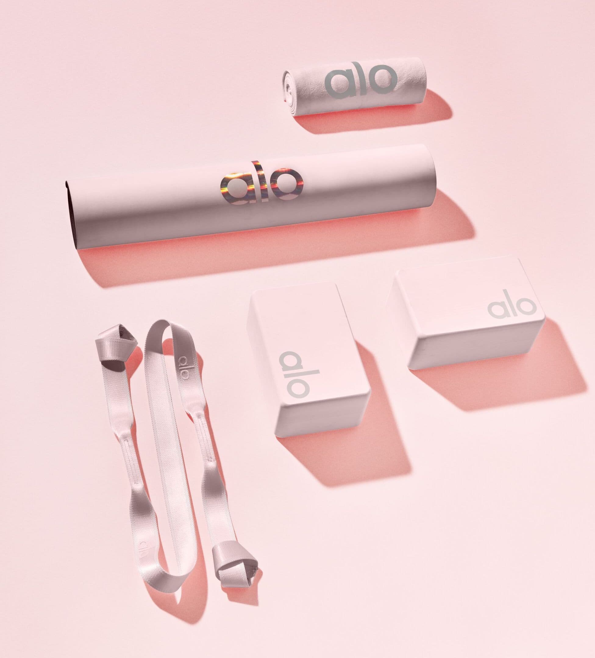 A photo of an Alo yoga mat, yoga strap, yoga blocks, and yoga non-slip mat in a light pink color displayed on a light pink background.  