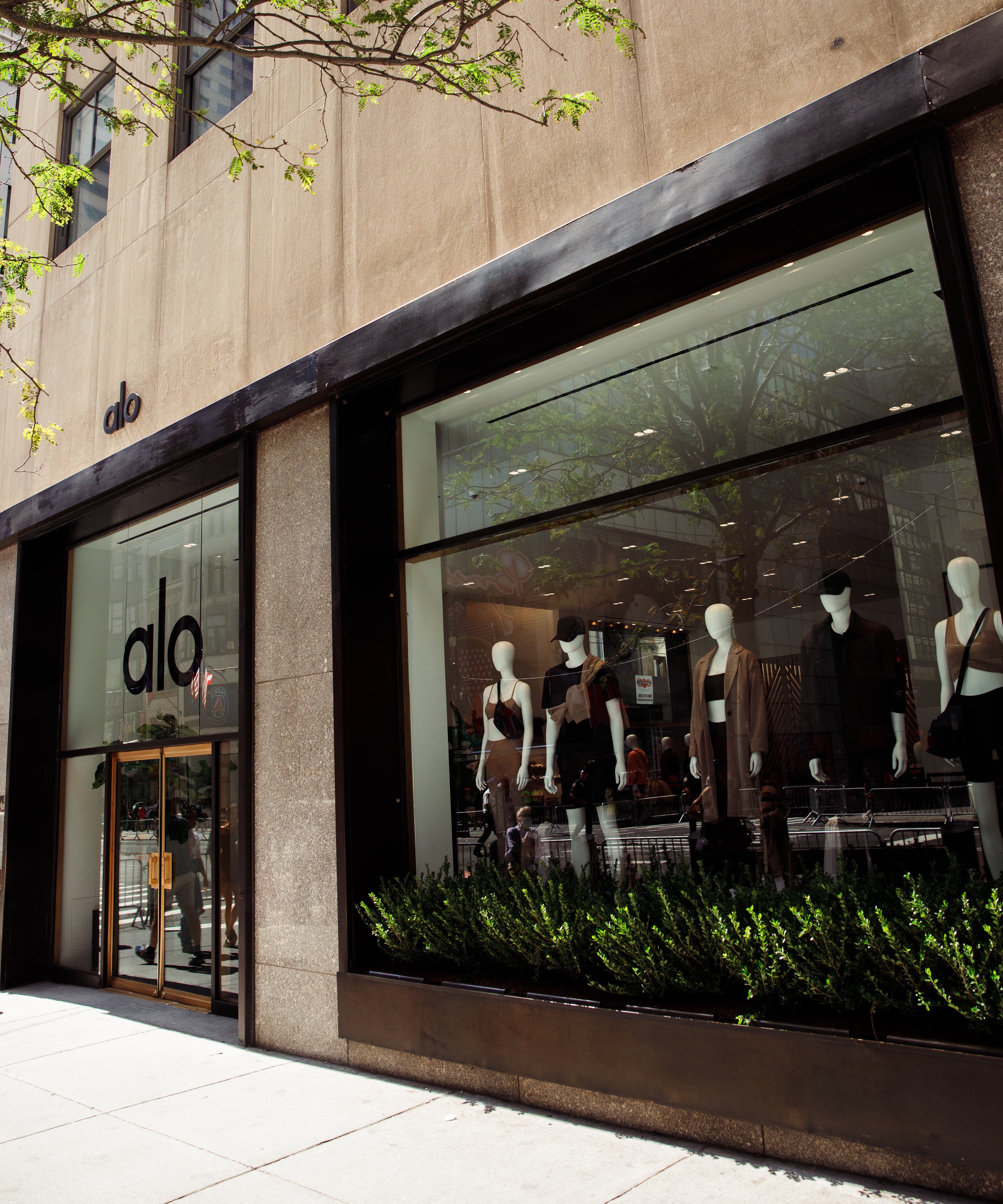 A photo of the Alo Yoga storefront at the Rockefeller Center in New York City featuring matte black framing of the windows with mannequins displayed wearing the newest Cinnamon Brown color paired with black. 
