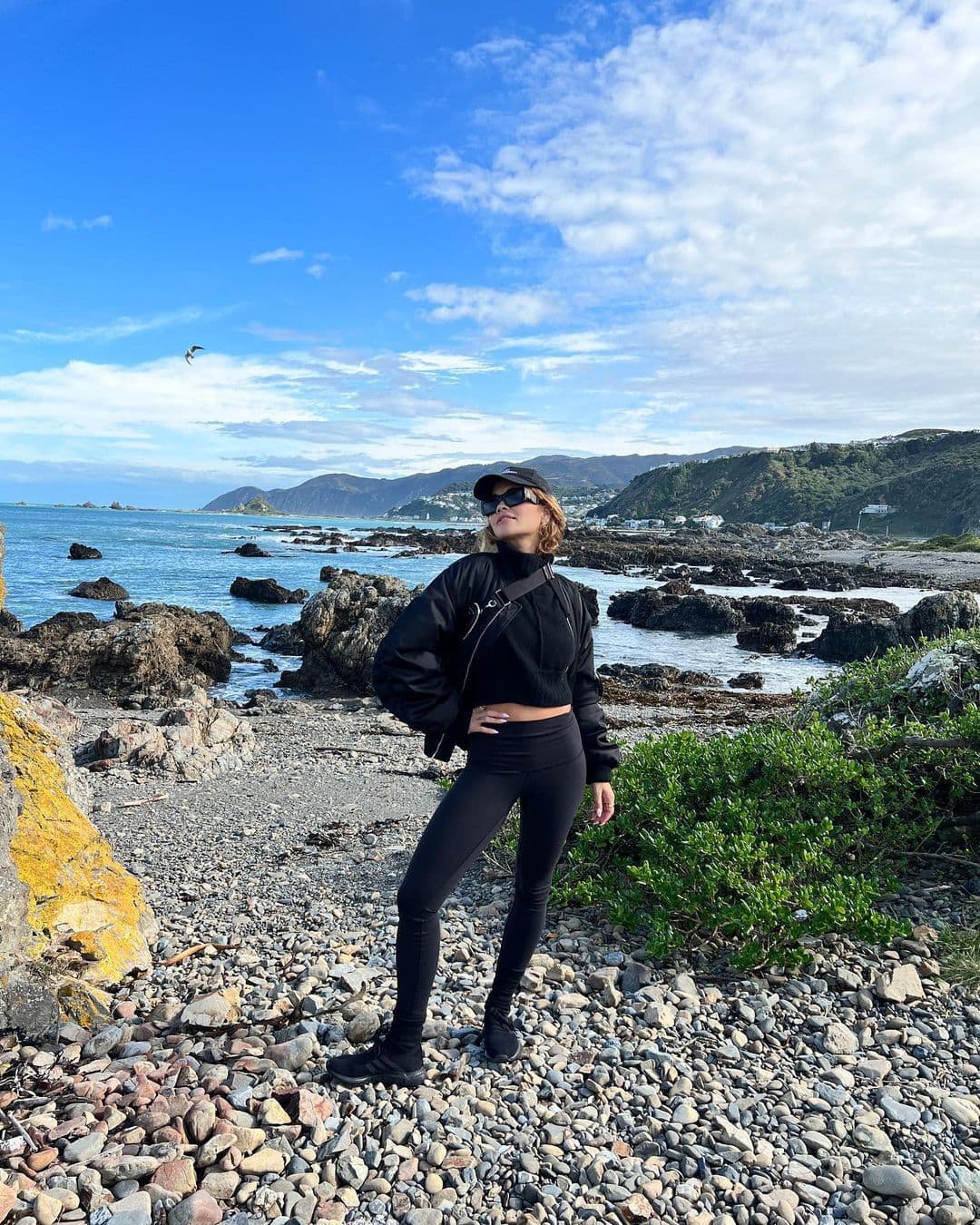 Rita Ora wearing a pair of black Alo leggings with a black bomber, sunglasses, and hat while standing on a rocky beach shoreline.  
