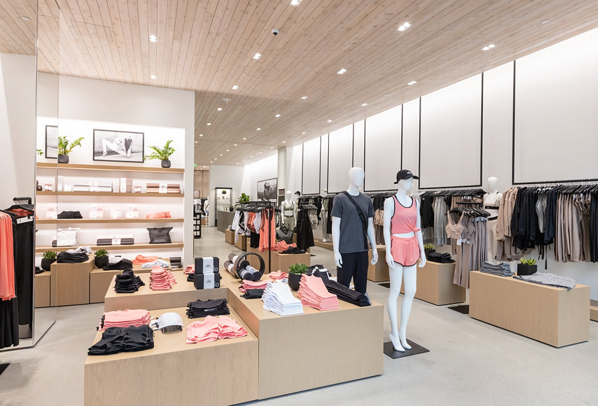 A photo of the inside of an Alo Yoga store with natural wooden blocks and mannequins displaying apparel with matte black modern hanger rods also showcasing the newest Alo Collections in the back. 