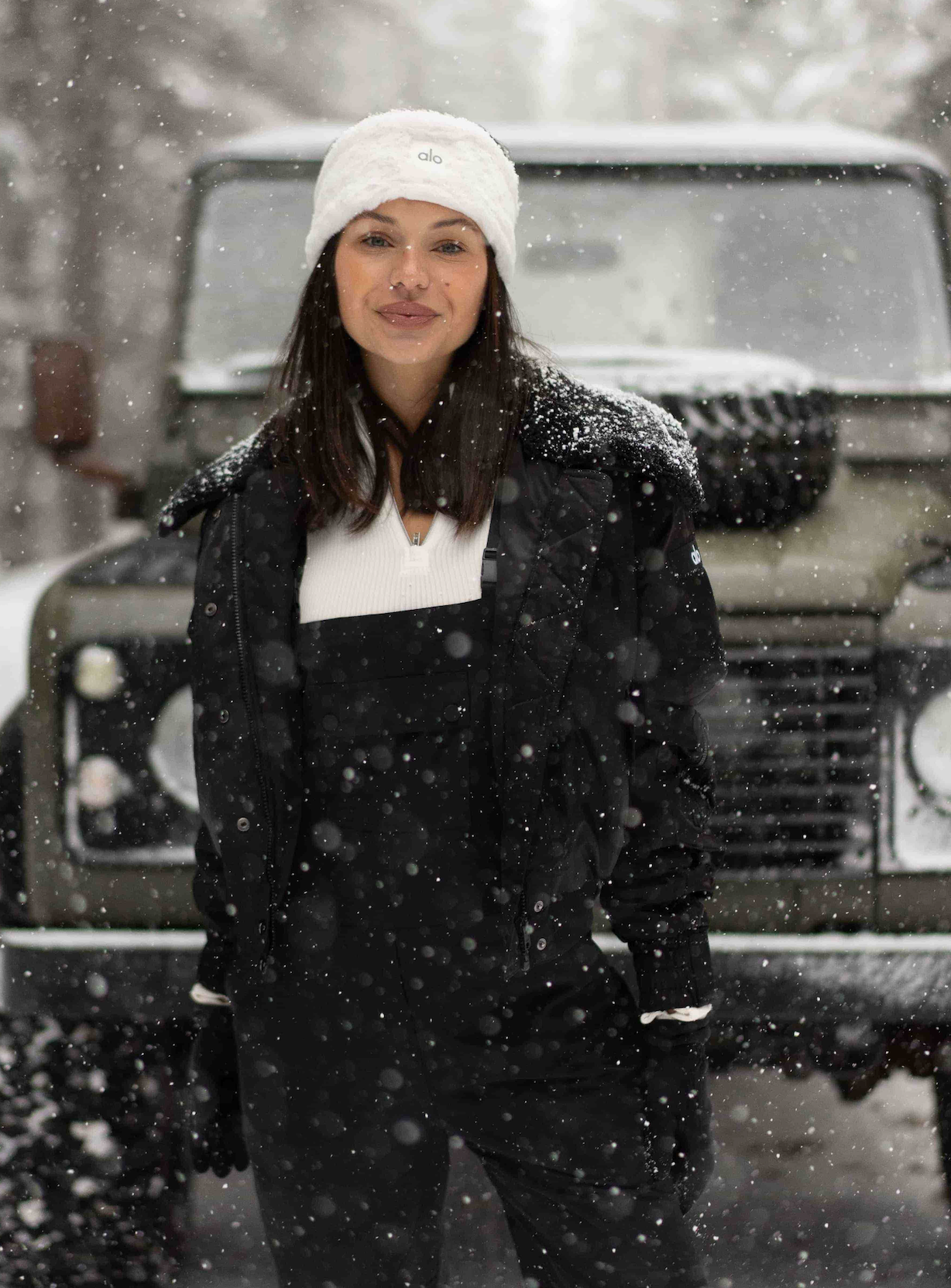 @alyssalynch wearing a pair of winter overalls, a long sleeve bodysuit, and a sherpa-collared jacket with a white faux fur headband standing in front of a large jeep in a snow-covered road with snowy pines lining the way. 