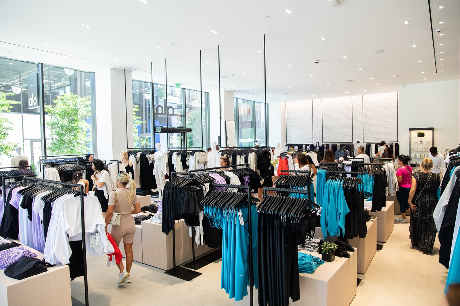 A photo of the inside of the Alo Boston Sanctuary showcasing many shoppers looking through various colored activewear in a large, naturally lit space.  
