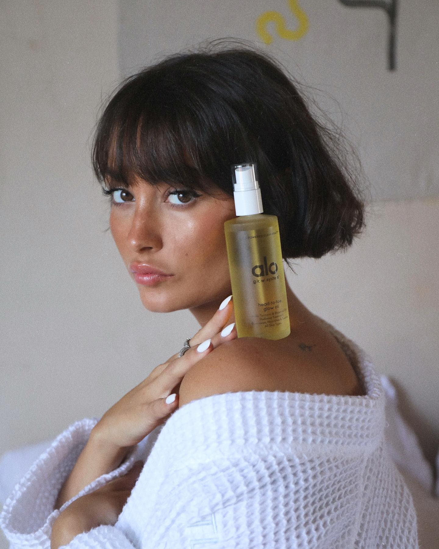 Woman with tanned skin, jaw length brown hair and curtain bangs poses with Head-To-Toe-Glow Oil bottle resting on her shoulder. 