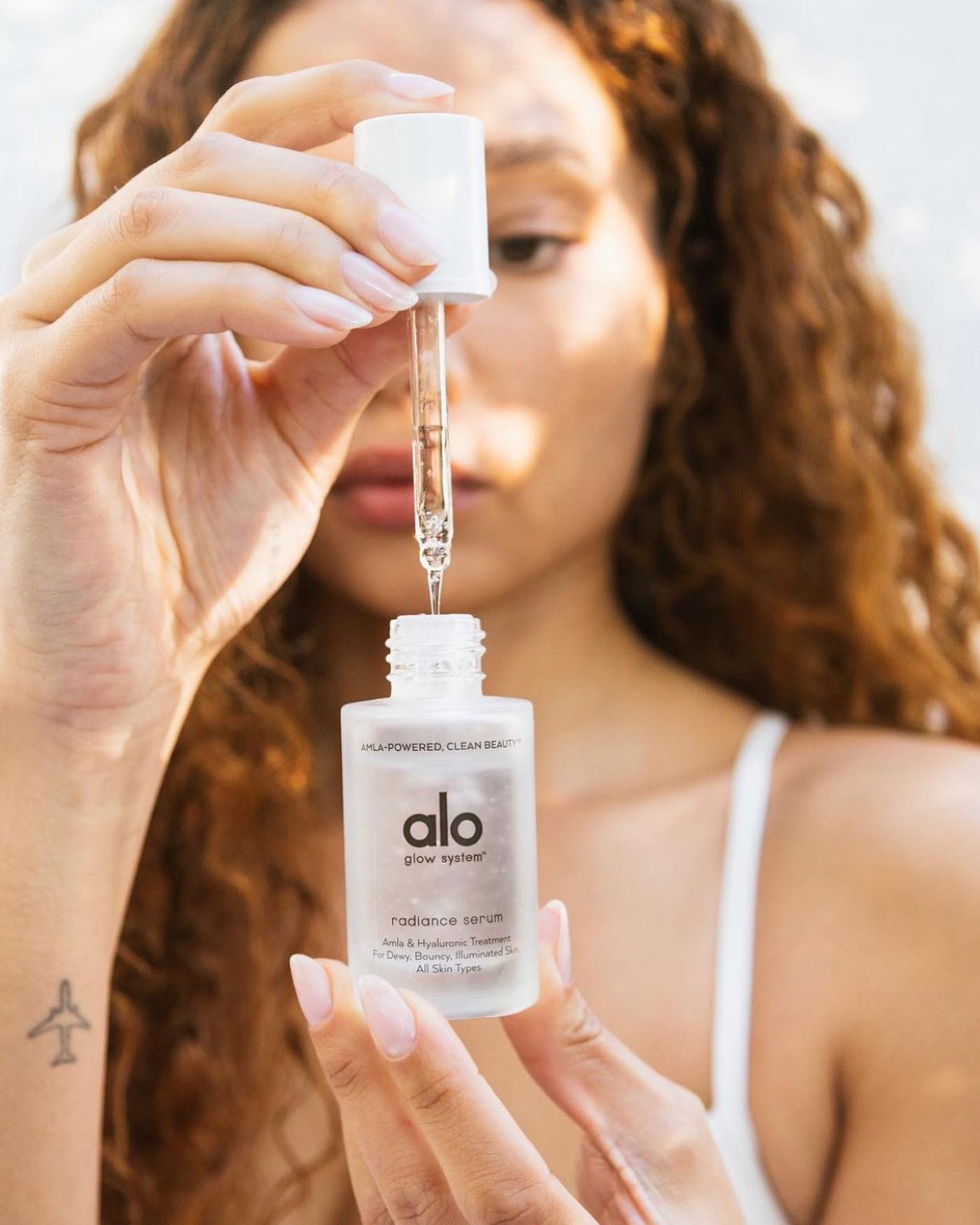 Close up on hands holding Radiance Serum dropper and bottle in foreground, woman with curly red hair holding the bottle in background 