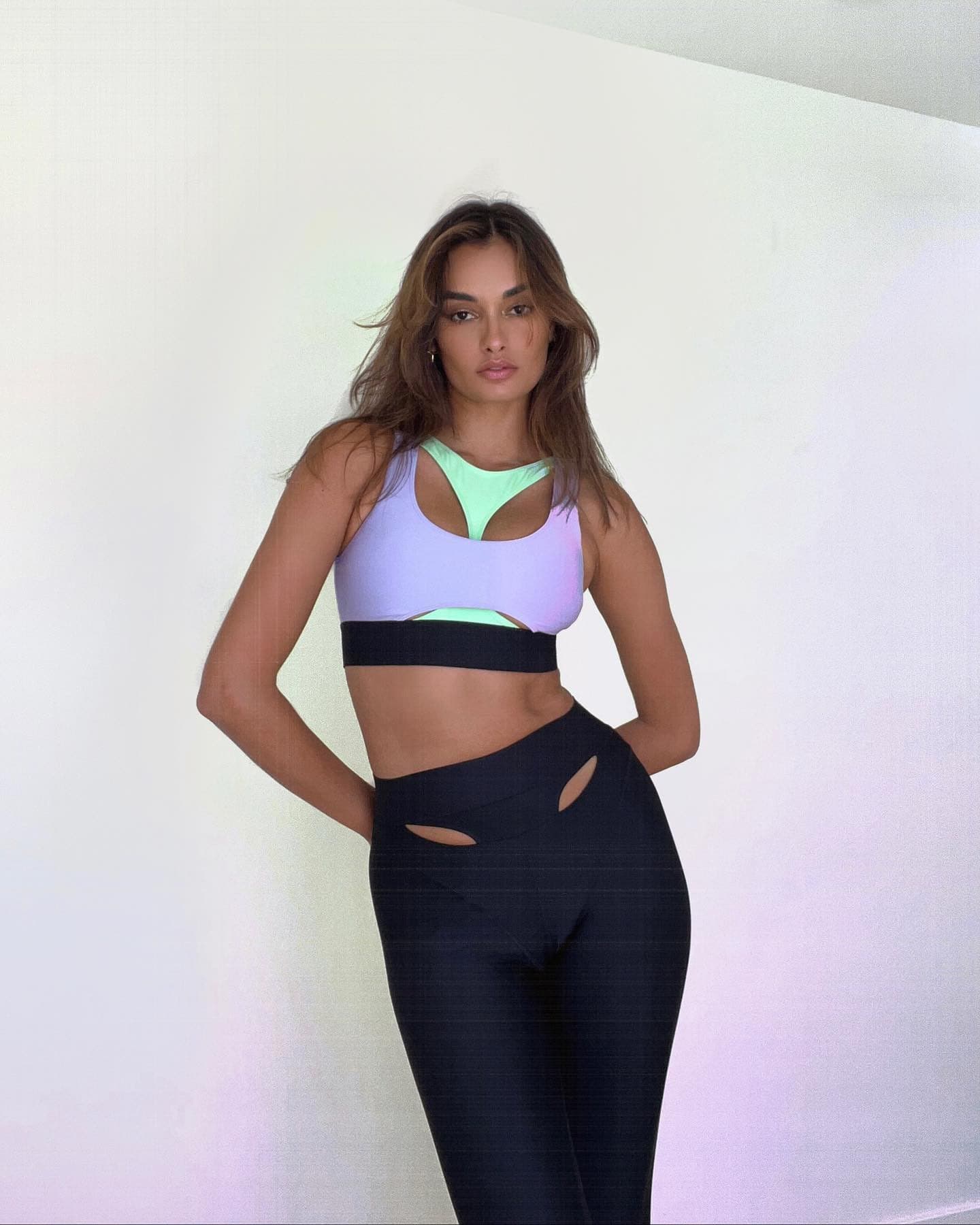 @giizeleoliveira wearing a neon purple and green bra with statement cutouts paired with a pair of black leggings featuring front high-waist cutouts while posing against a white wall.  