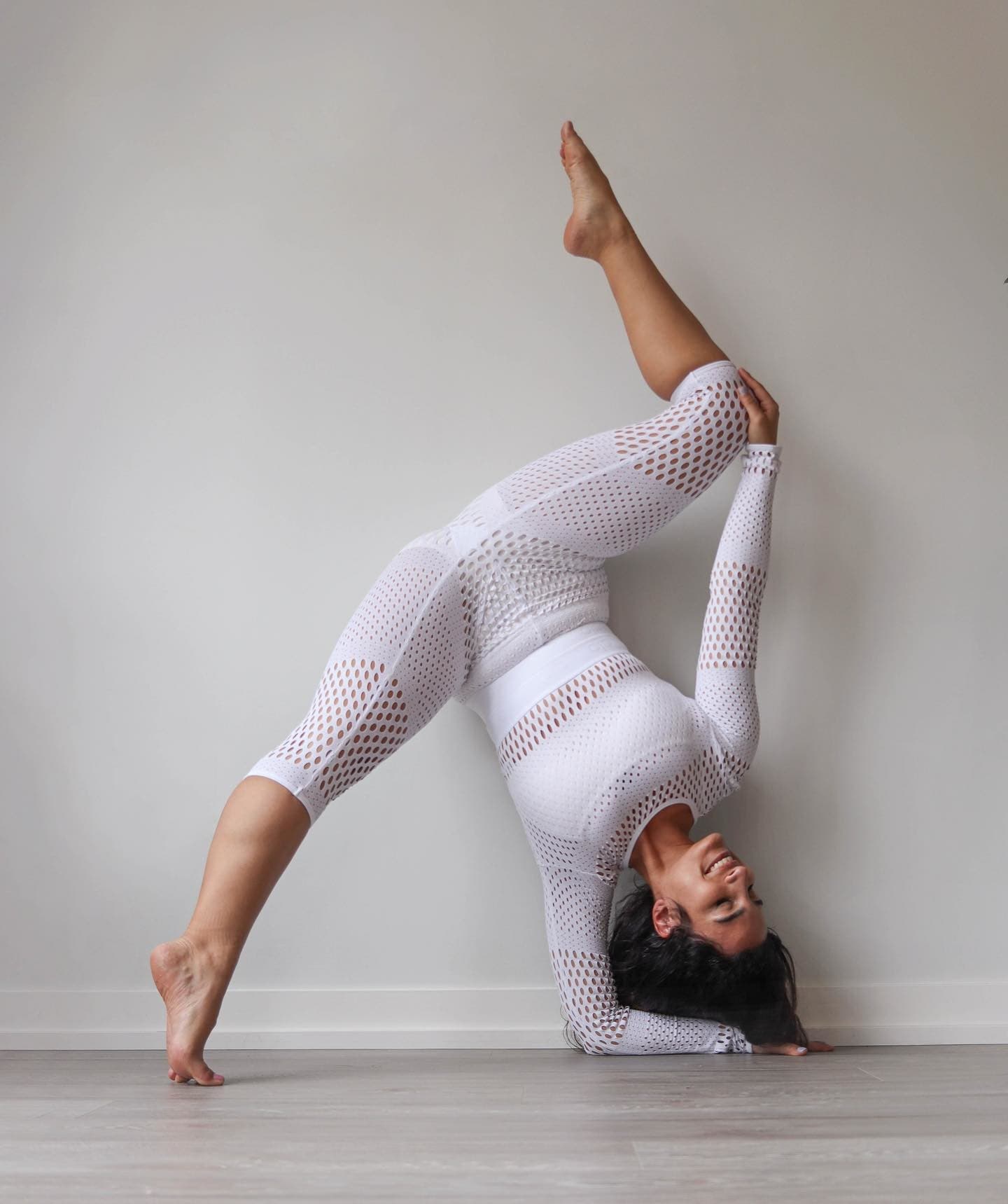 @asanavanessa wearing a Haute Mesh Summer Long Sleeve in White with a matching pair of Mesh High Waist Haute Summer Capris while posing against a white wall in a single forearm stand.  