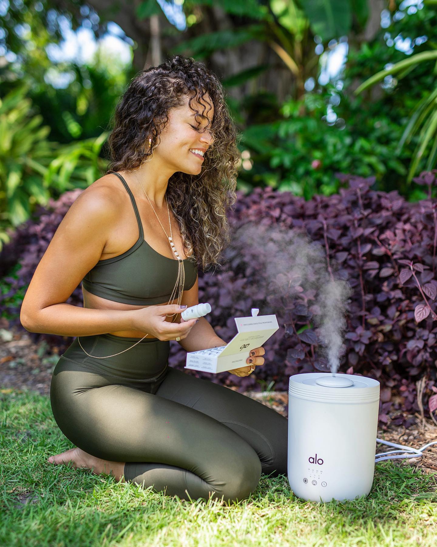 @bloggerswhobrunch post of woman on grass with curly hair, wearing an Alo Yoga Airlift Intrigue Bra and Leggings enjoying an Aura Essential Oil Diffuser 