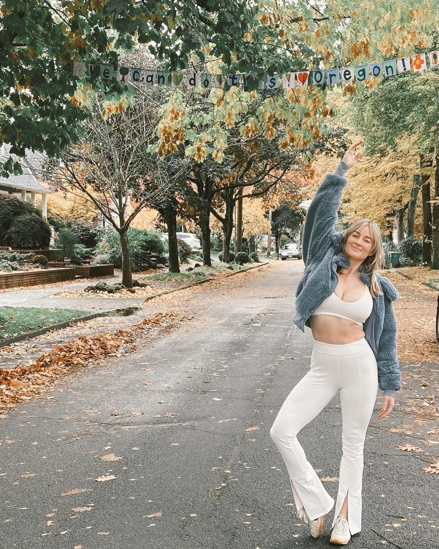 @caleyalyssa wearing a pair of flare leggings with fronts slits and a matching sports bra in ivory with a plush sherpa jacket while posing in a street with colored leaves lining the sidewalks and yellow and orange trees lining the streets. 