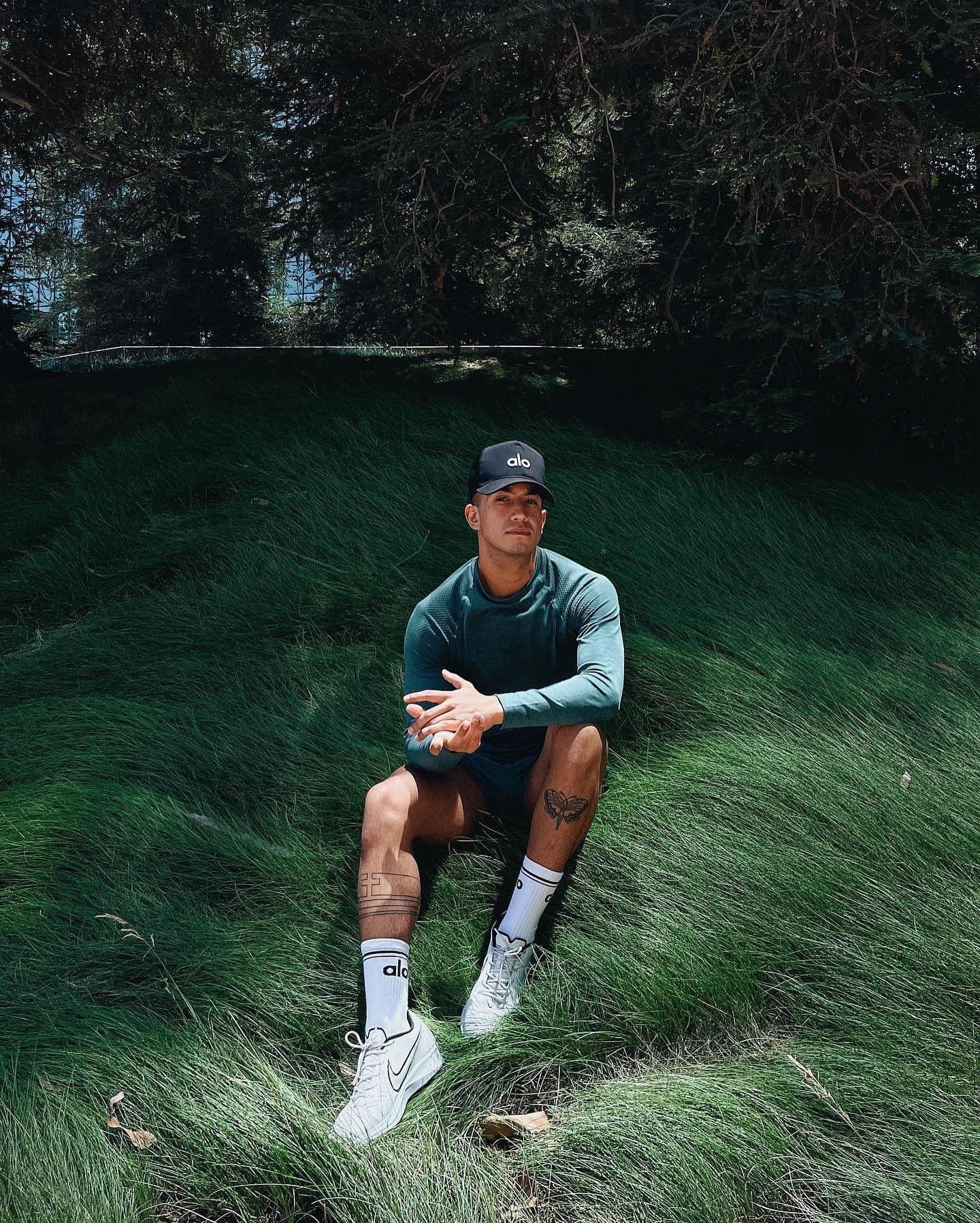@christiancaro_ wearing a dark teal hooded runner jacket with a pair of Alo crew socks and a black Alo trucker hat while sitting in a patch of long grass.  
