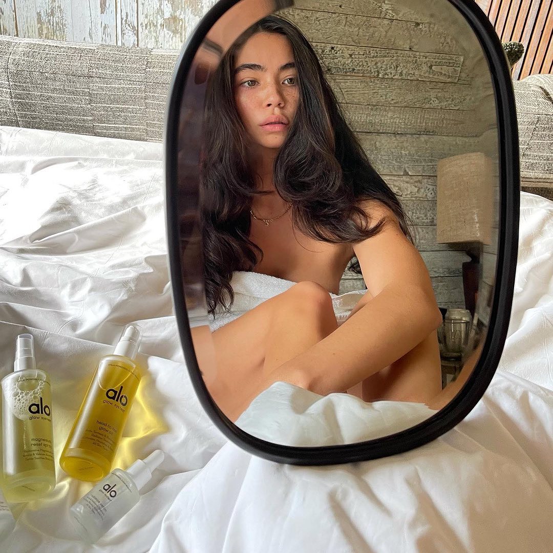 @credobeauty girl with long dark sits on a bed with Radiance Serum, Head-To-Toe-Glow Oil, and magnesium Reset Spray bottles while looking into a mirror. 