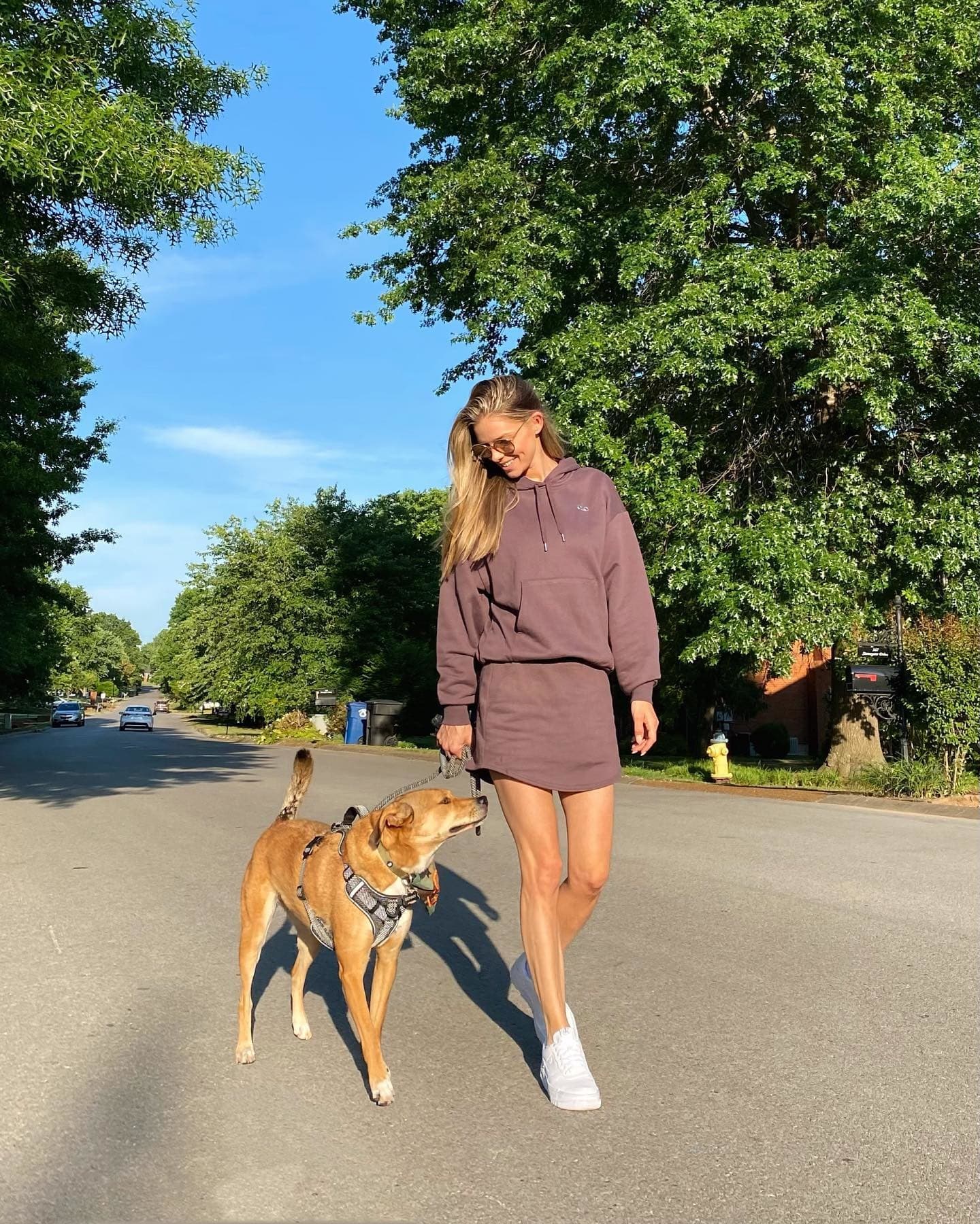 @danielleknudson1 porte une jupe en sweat marron avec un sweat-shirt à capuche assorti alors qu'elle promène son chien dans une rue bordée de grands arbres verts.