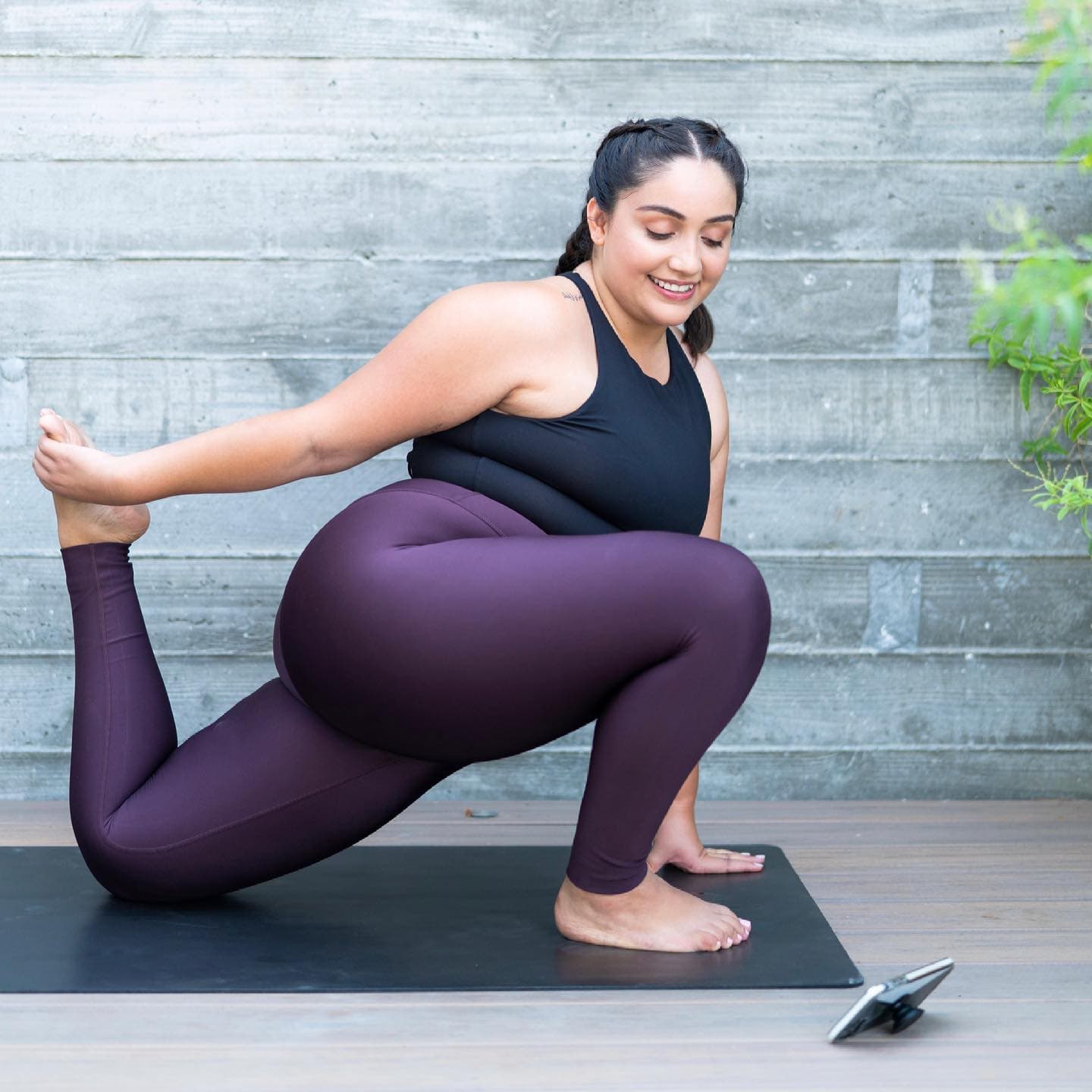 A woman stretching on her black yoga mat using the Alo Moves App while wearing a pair of dark purple high-waisted leggings and a black high-neck workout top. 