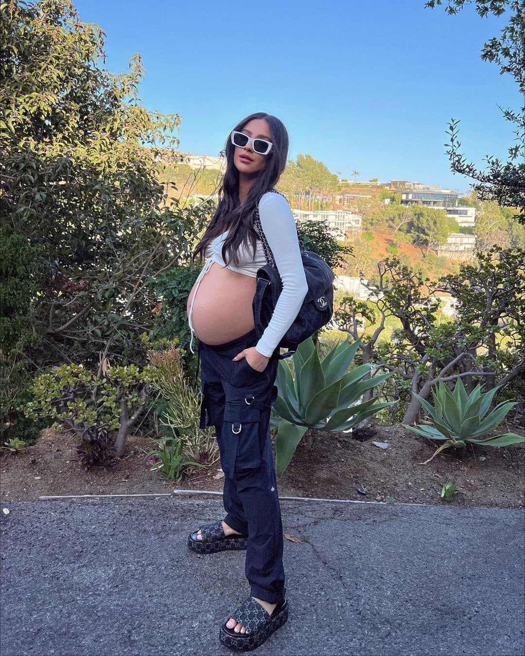 A photo of Shay Mitchell wearing a pair of black Alo cargo pants with a long sleeve front cinch top while posing on a road overlooking houses in the background.  