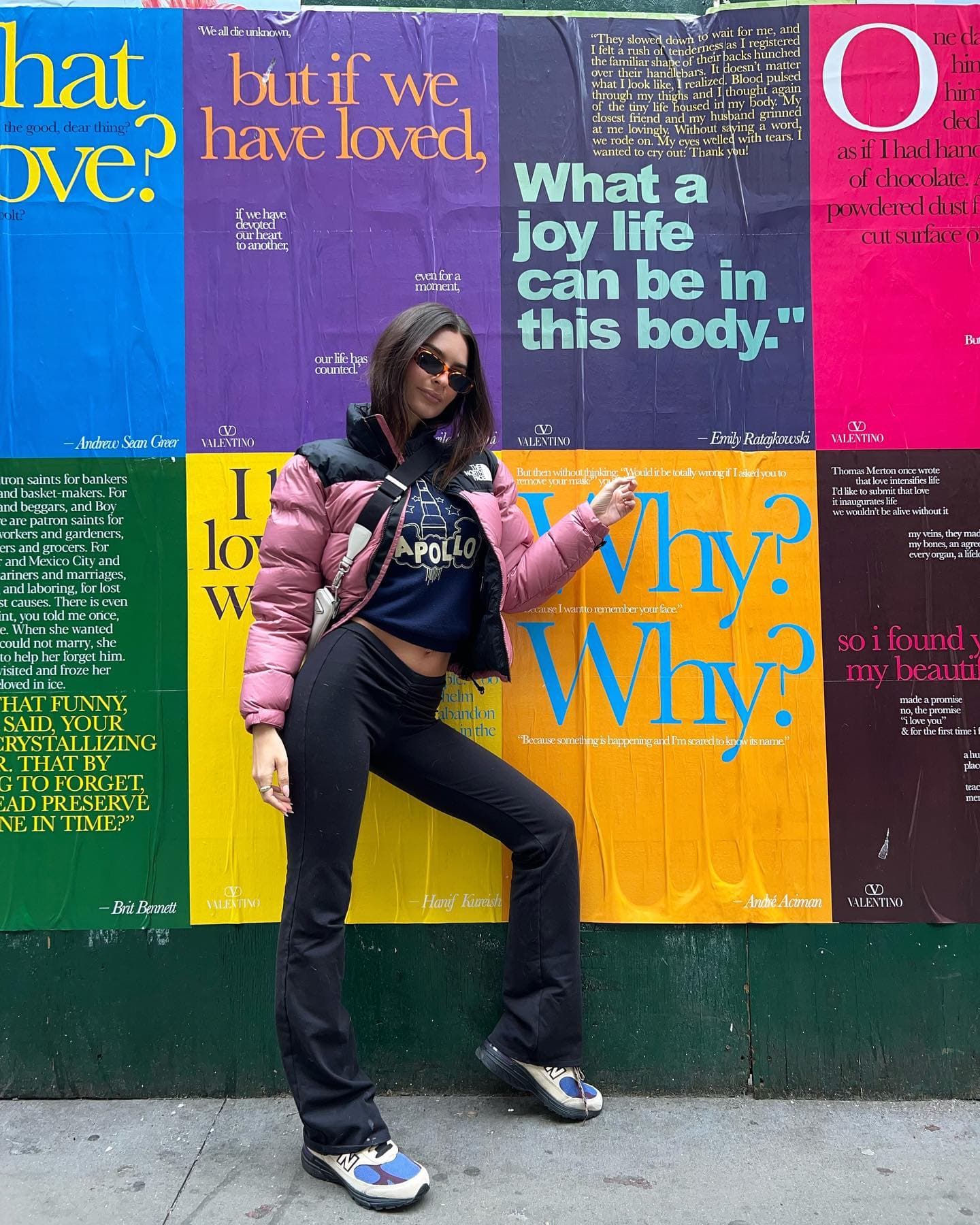 A photo of Emily Ratajkowski posing in front of a wall of posters while walking around New York City.  