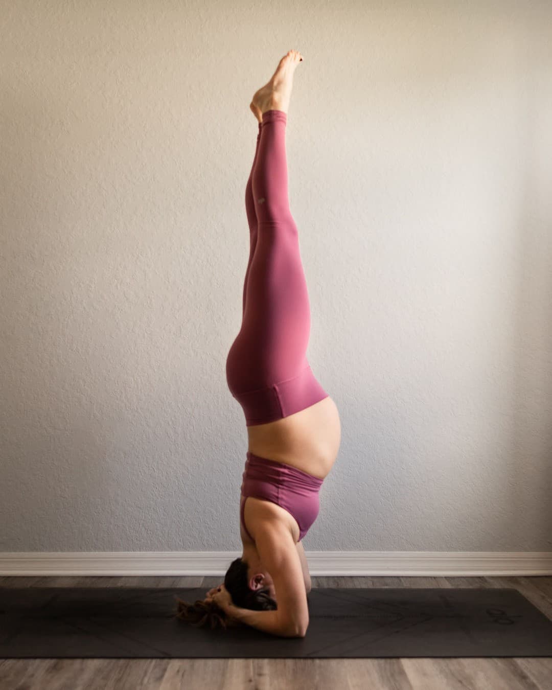 A woman wearing a violet-colored workout set with high-waisted leggings and a sports bra tank while in a forearm headstand while pregnant. 
