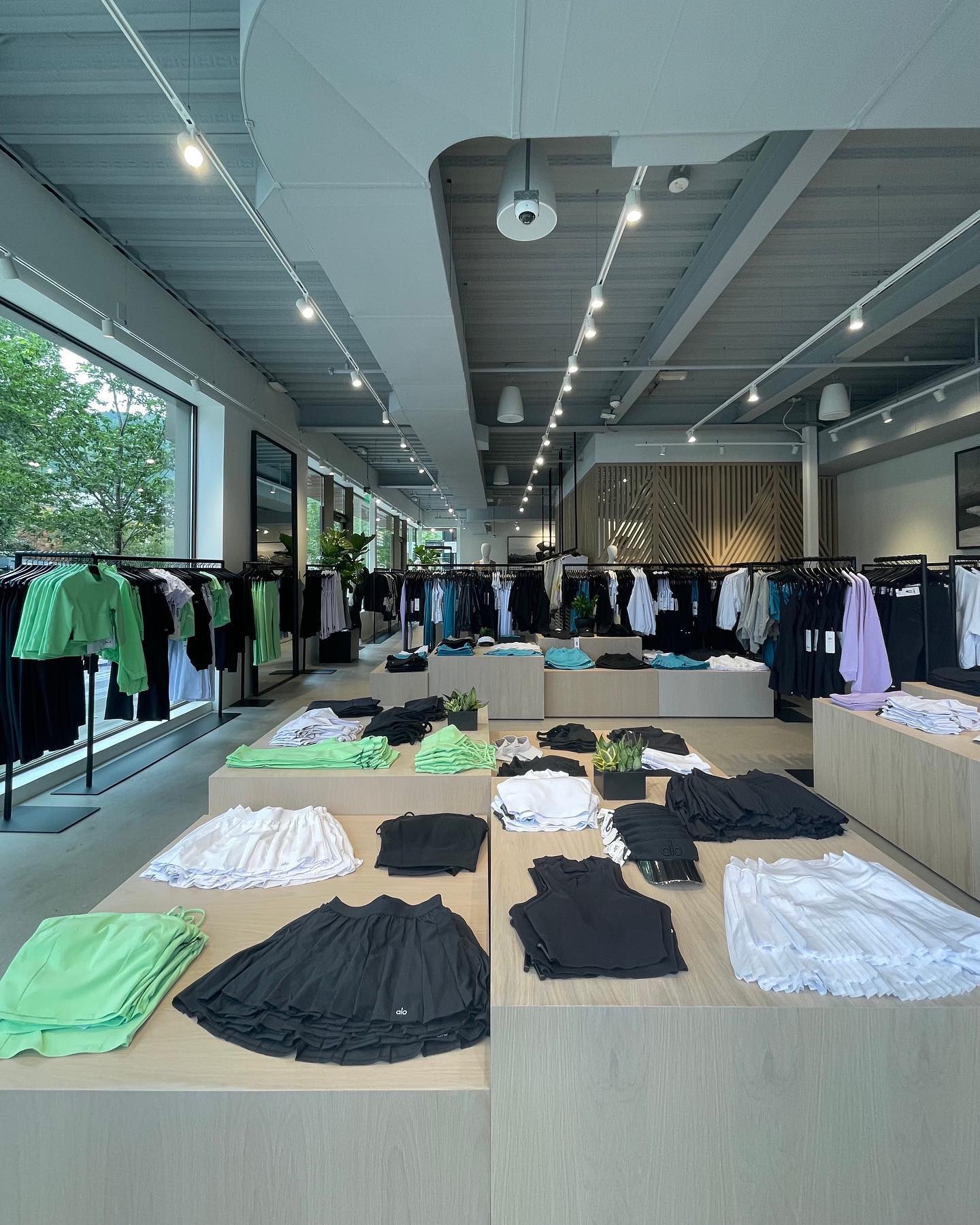 A photo inside Alo Aspen showcasing several large natural wood boxes with tennis skirts, workout tops, and leggings displayed on top with an array of matte black racks displaying activewear jackets and tops in the background.  