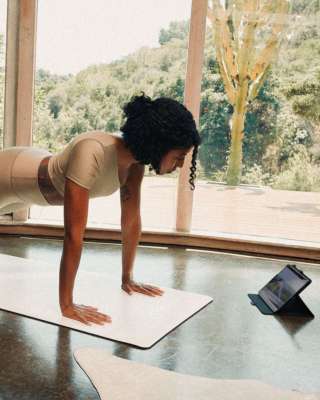 Woman in plank position doing Alo Moves from an iPad on the floor in front of her with a large window behind her showing a desert greenery and cactus landscape. 