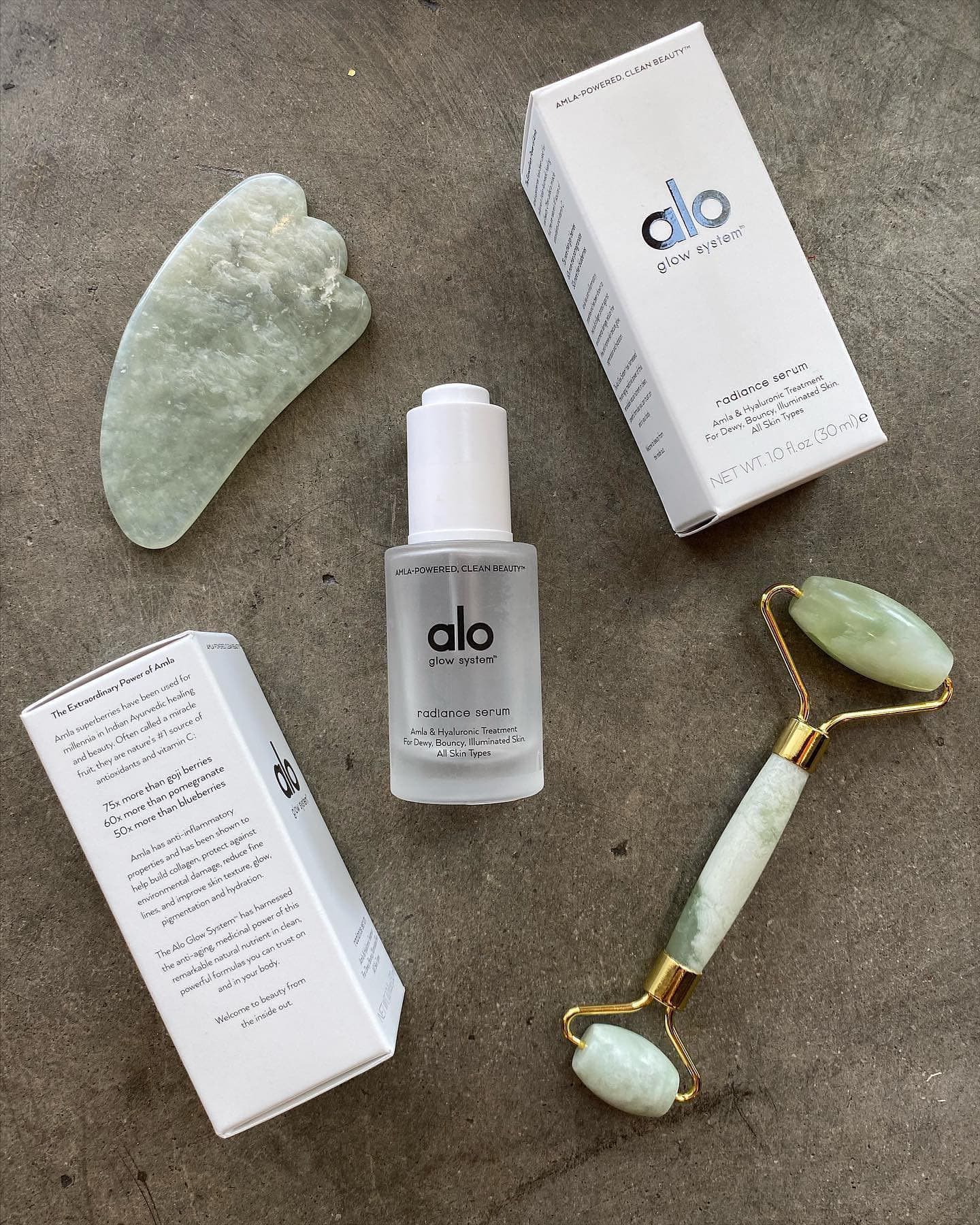 A flatlay photo on a concrete surface of Alo’s Radiance Serum surrounded by a jade roller and gua sha tool.  