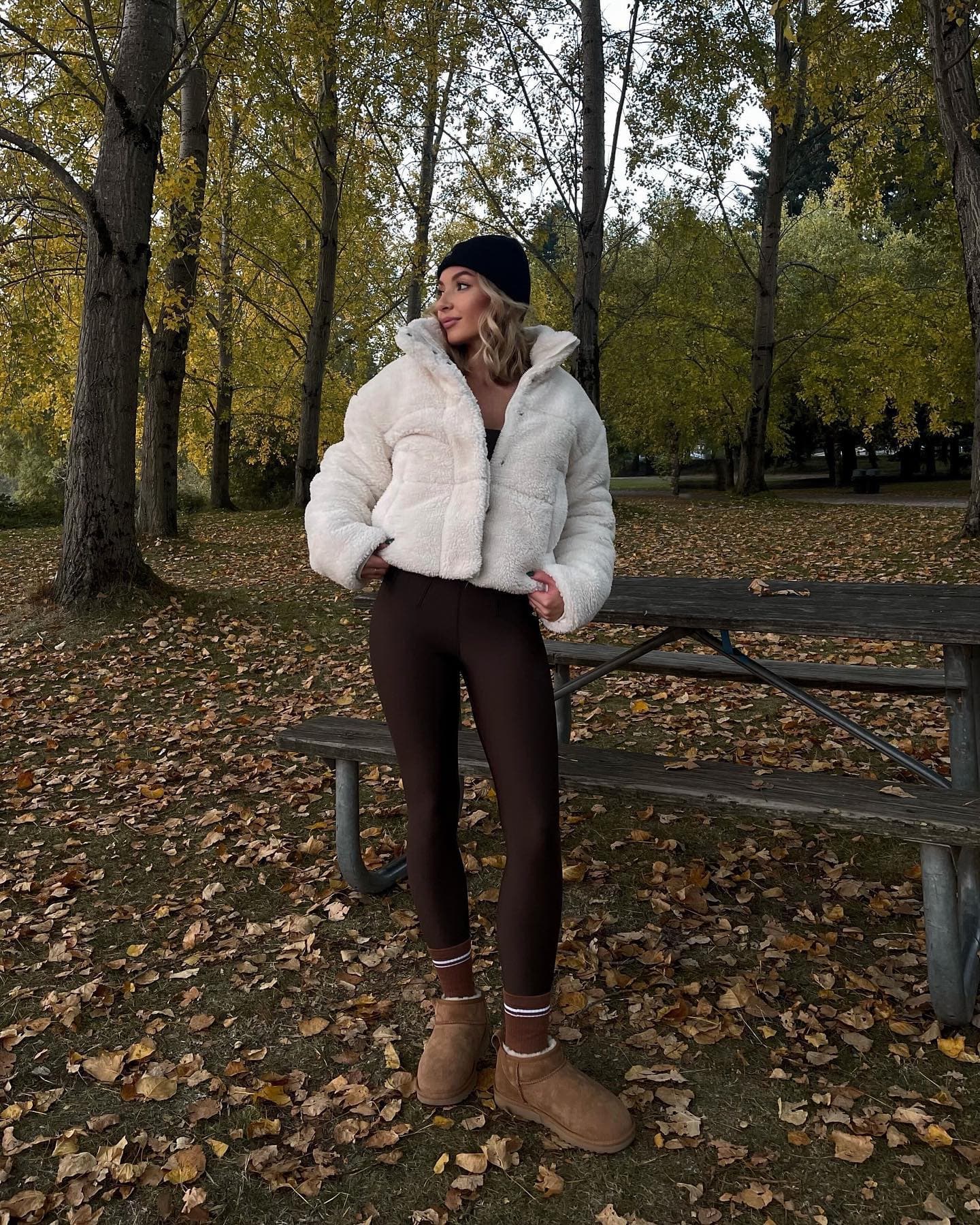 @gingeriley wearing an ivory sherpa puffer with a pair of brown high-waisted leggings, brown Alo socks, a black beanie, and a pair of boots while standing in an empty park with lots of leaves on the ground.  