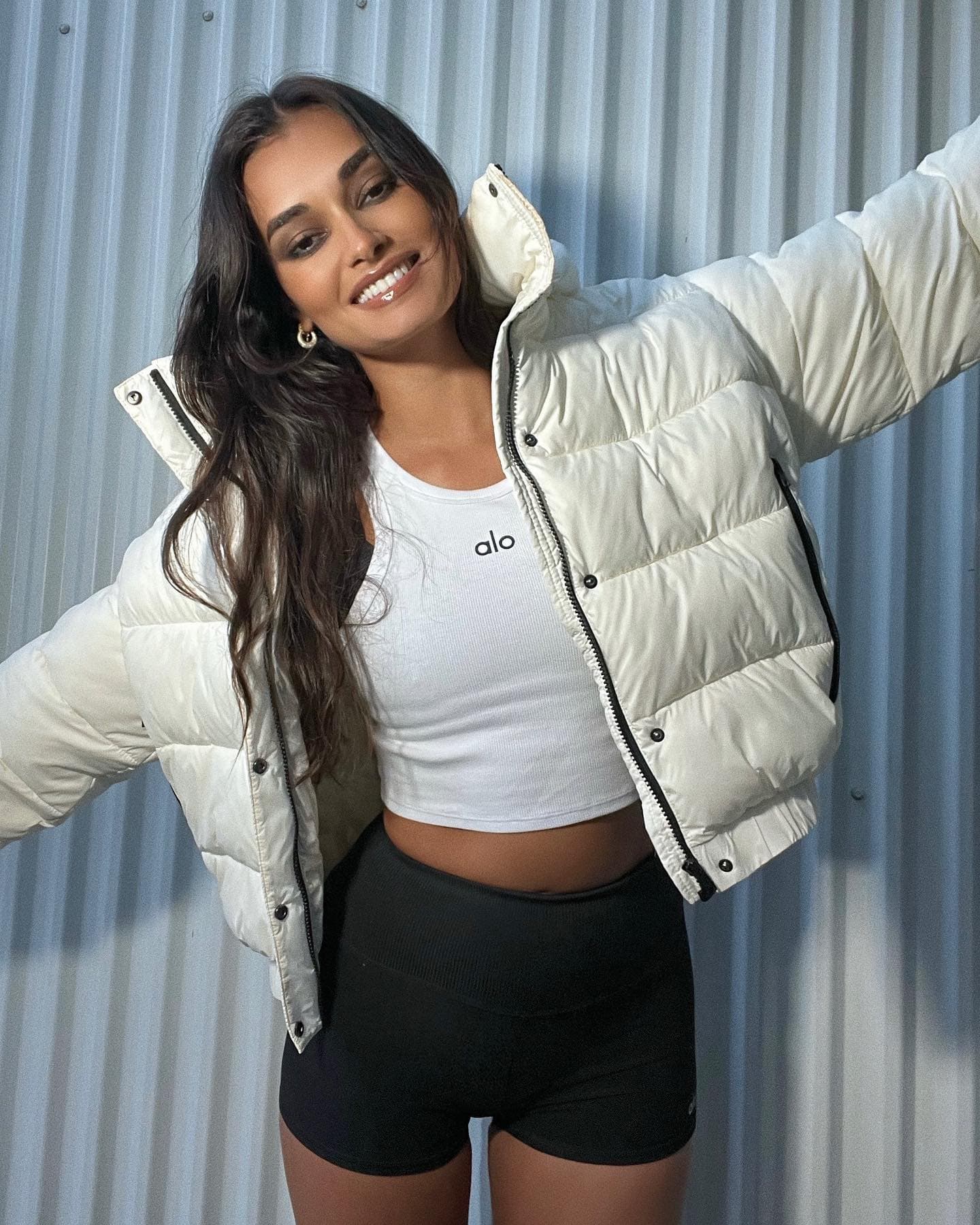 @giizeleoliveira wearing a white puffer jacket with black contrast piping paired with a white Alo tank top and black shorts while standing in front of a white wall. 