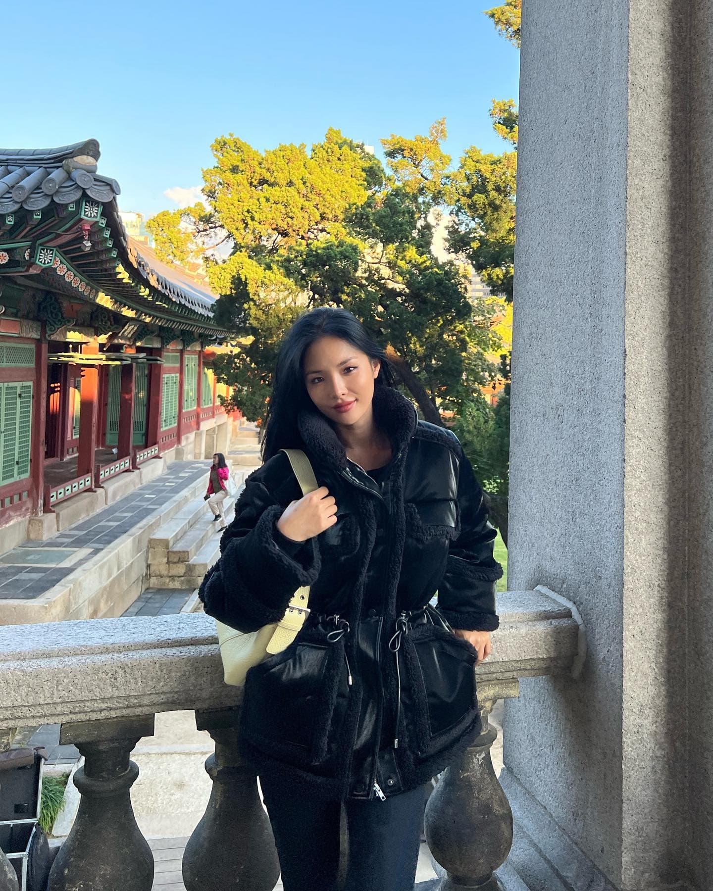 @chaileeson wearing a black satin faux leather coat with sherpa details and a longer length paired with black leggings while standing in front of a temple on a balcony overlook. 