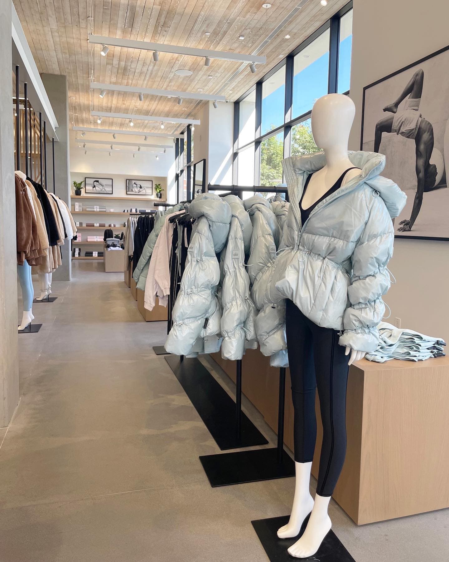 A photo of the interior of an Alo Yoga store displaying mannequins wearing winter coats and natural wood blocks with folded apparel on top. 