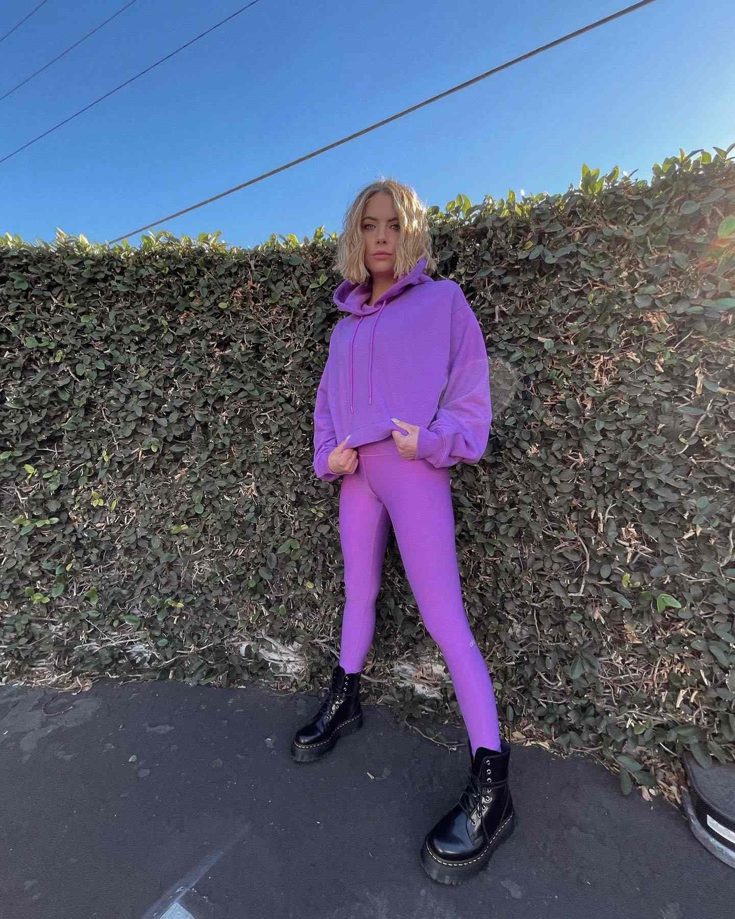 Ashley Benson porte des leggings Airlift taille haute Electric Violet et un sweat à capuche Electric Violet Bae assorti avec de grosses bottes noires tout en posant contre un mur de béton recouvert de lierre.