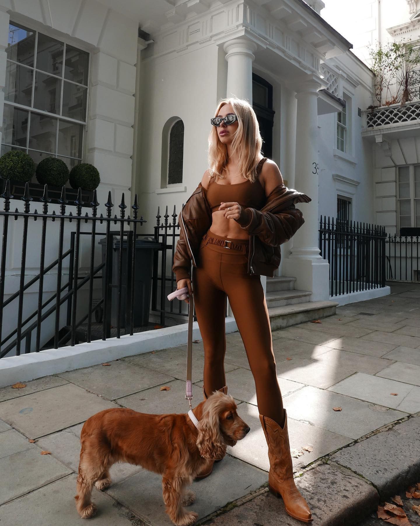 @kimberleygarner wearing matching brown workout set with high-waisted leggings and a sports bra featuring scrunched strap and belt details with a pair of cowboy boots and a faux leather bomber jacket while walking her dog on a sidewalk.  