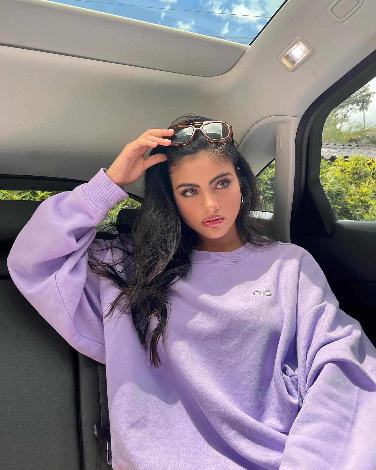 @saraorrego wearing a neon purple crew neck sweatshirt while sitting in a car posing with her hand on her sunglasses that are propped on the top of her head.  