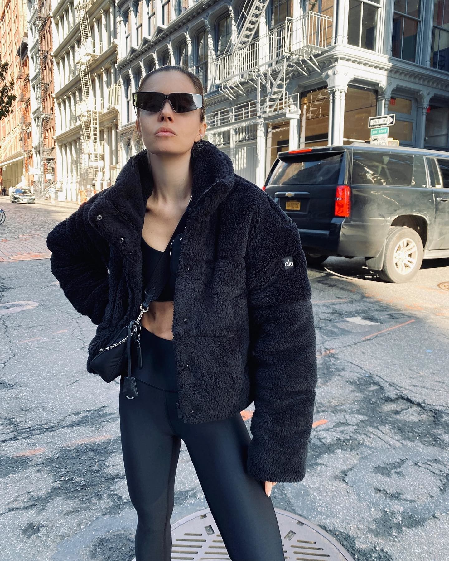 @elizabethsulcer wearing a black sherpa puffer jacket with a pair of black high-waisted leggings and a matching black sports bra while standing in a street in New York City. 