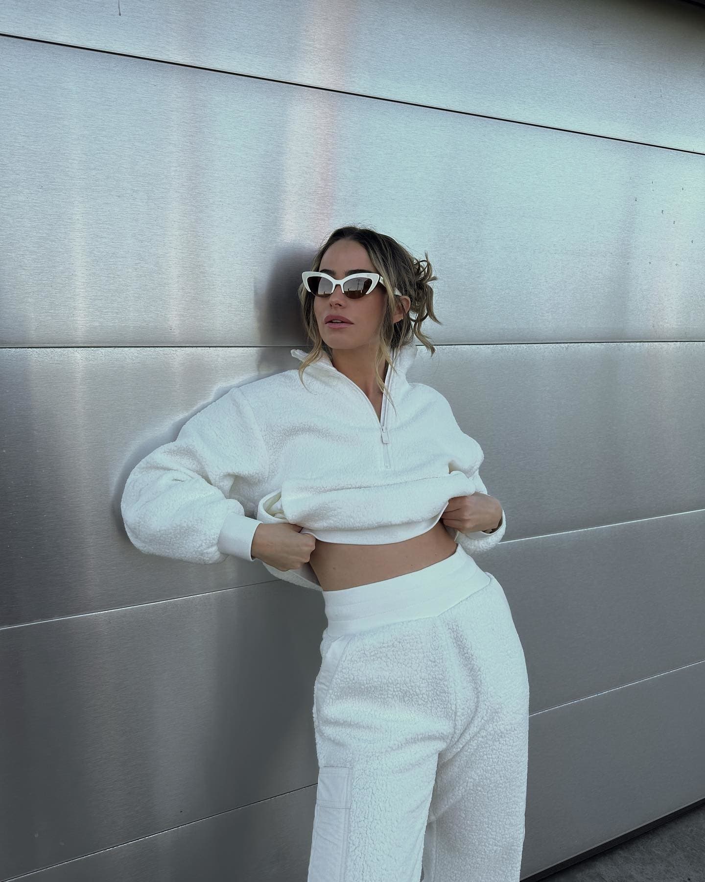 @ewebzz wearing a micro sherpa quarter zip jacket with a matching pair of micro sherpa sweatpants with cat-eye sunglasses standing against a silver metal wall.  