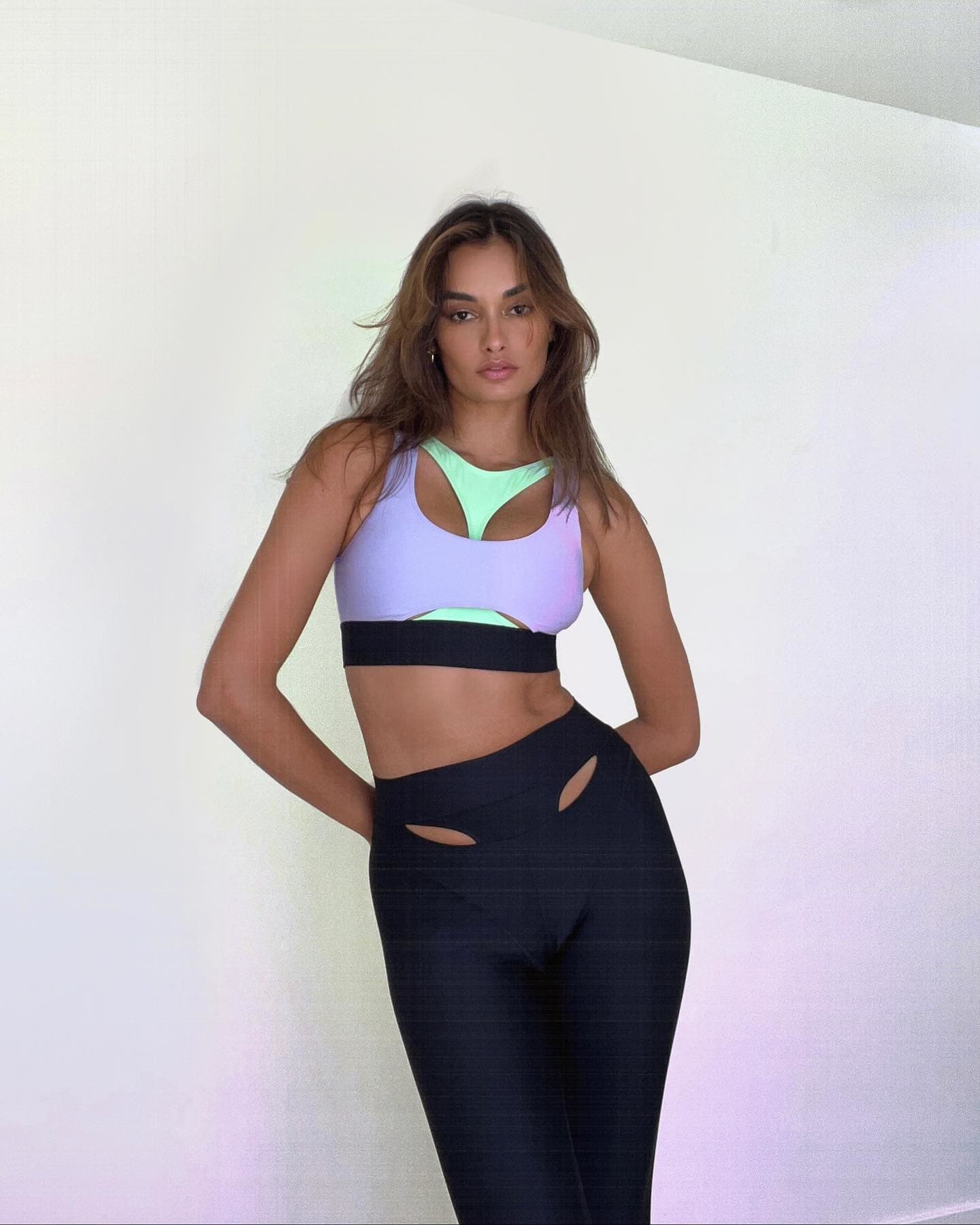 @giizeleoliveira wearing a purple and green statement sports bra with a pair of black statement workout leggings while posing with her arms behind her back in front of a white wall. 