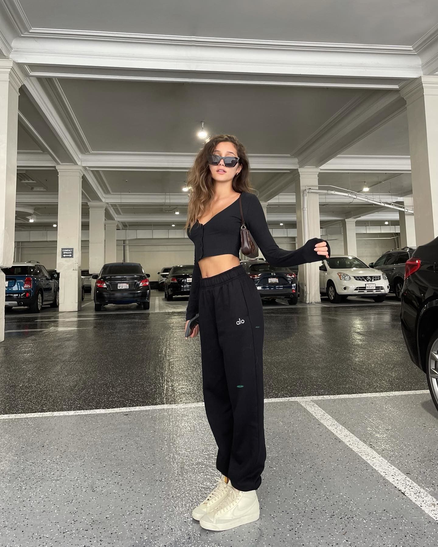 @haileysani wearing a pair of black Alo sweatpants with a matching black cropped cardigan while standing in a parking lot looking at the camera. 