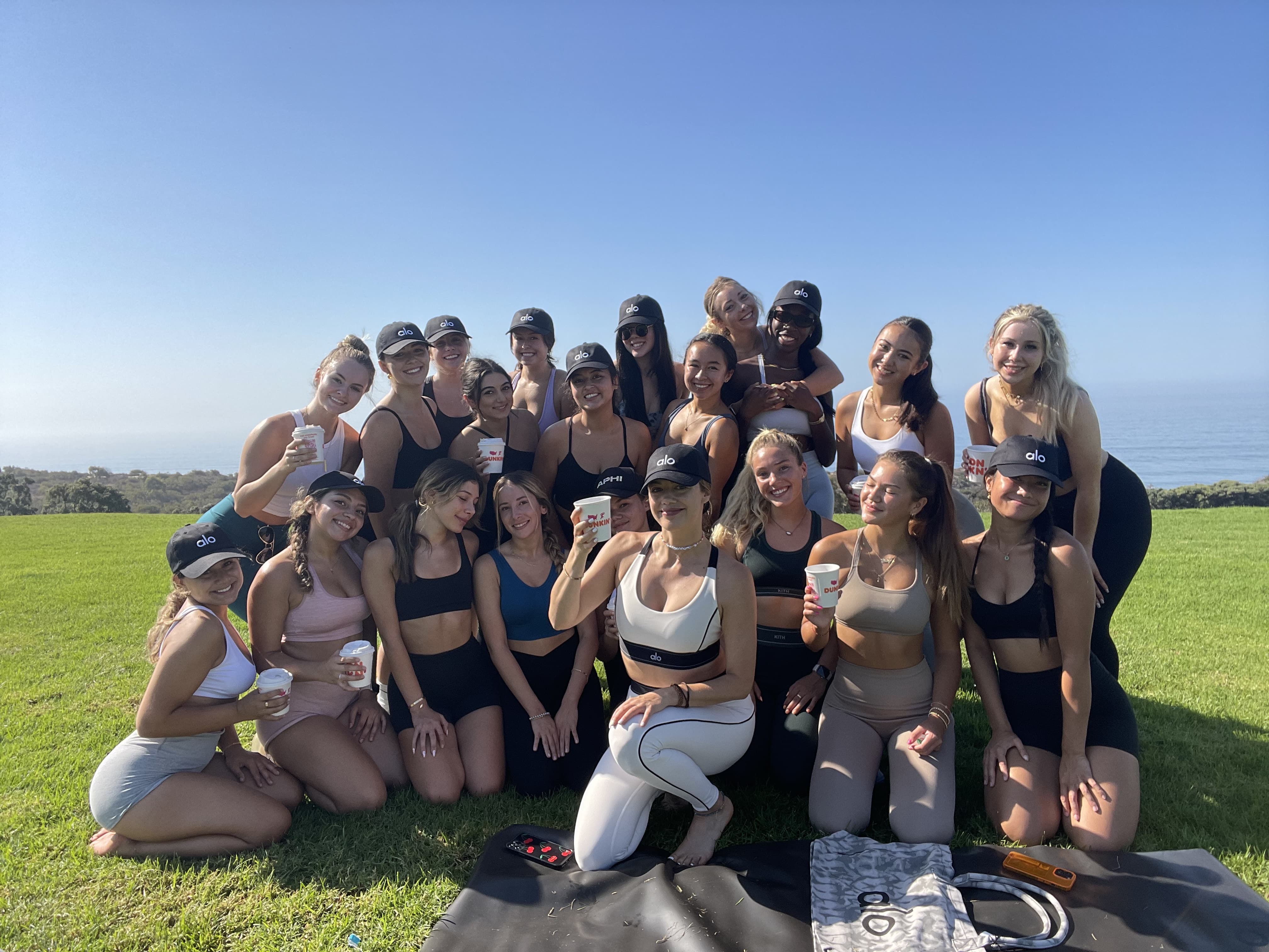 A photo of Britt Turpack and young women from Pepperdine posing for a group photo on a grassy hill overlooking the ocean after practicing yoga with Alo for National Yoga Month.  