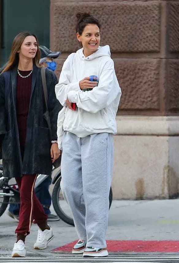 Katie Holmes wearing a pair of grey Alo straight leg sweatpants with a white oversized hoodie while walking along a crosswalk.  