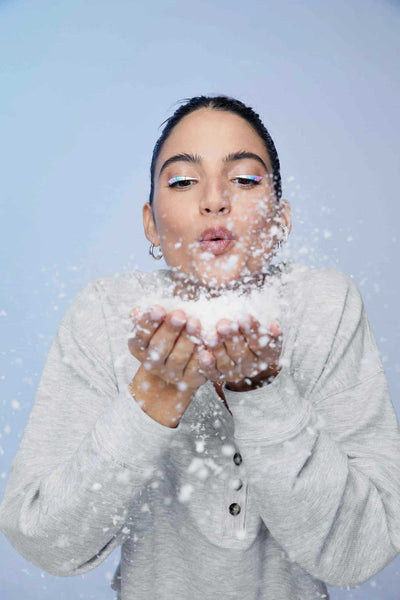 A woman blowing fake snow toward the camera while wearing a grey quarter button sweatshirt against a greyish blue backdrop. 