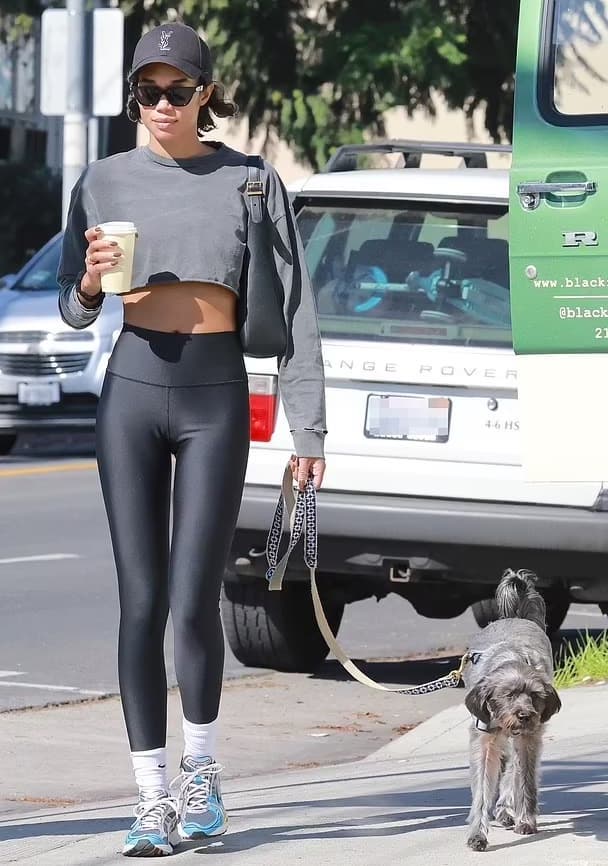 Laura Harrier wearing a pair of dark gray Alo leggings and a cropped long sleeve while walking her dog with coffee in hand along a sidewalk.  