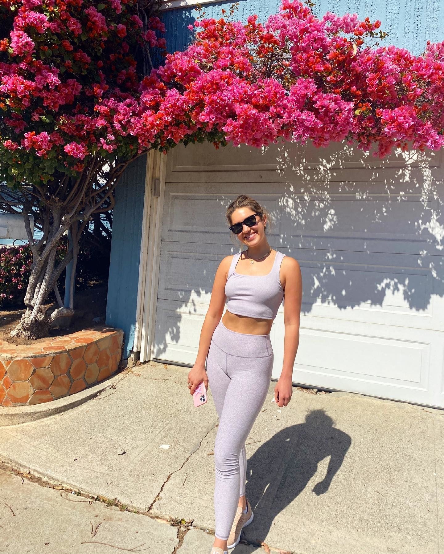 @Itsmetinx portant le Highlight Legging et le débardeur Alosoft Ribbed Chic Bra tout en posant près d'un arbre fleuri de fleurs magenta vives à l'extérieur d'une maison bleu royal.