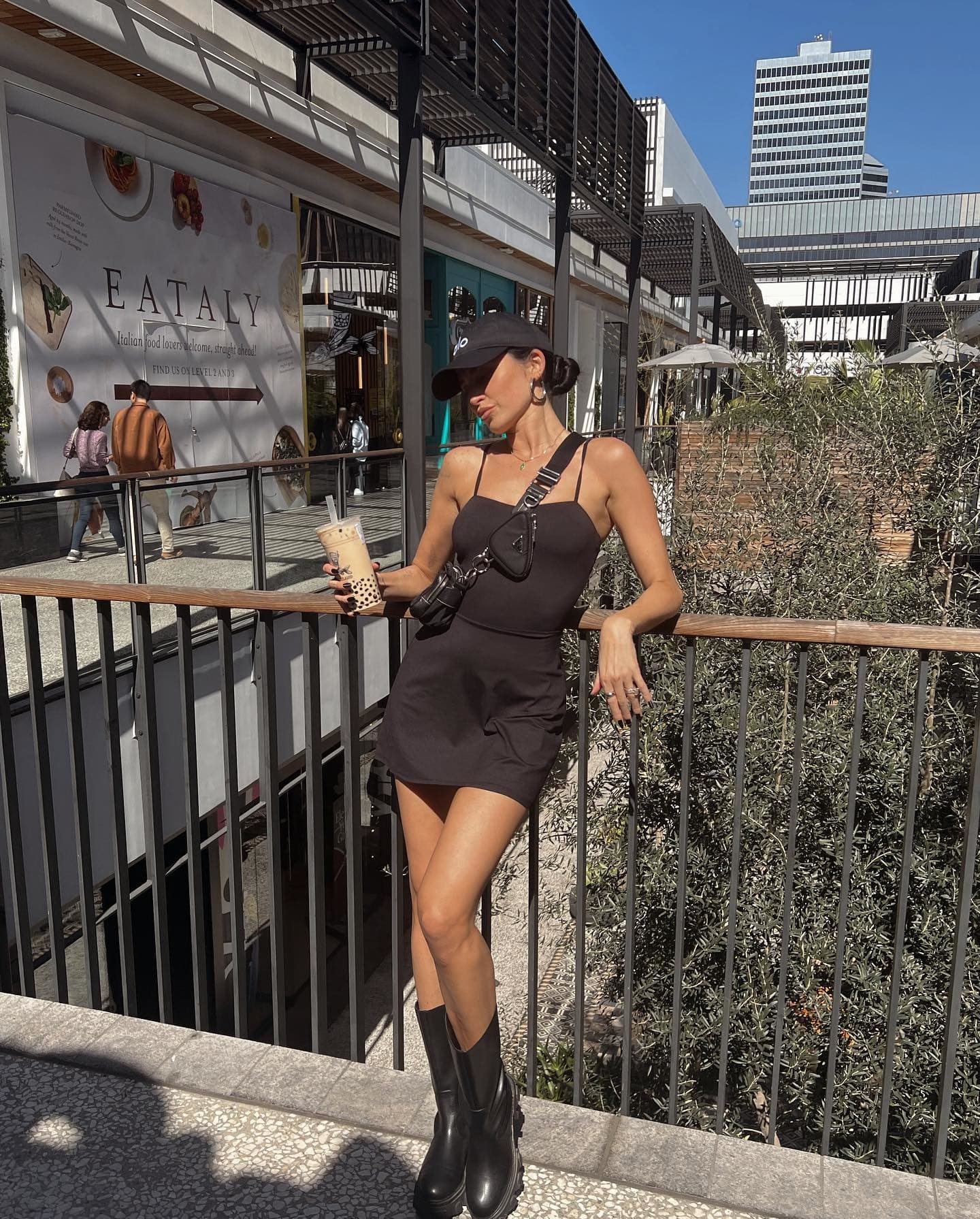 @justtnic wearing a black tennis dress with a black Alo hat while posing against a railing at an outdoor mall. 