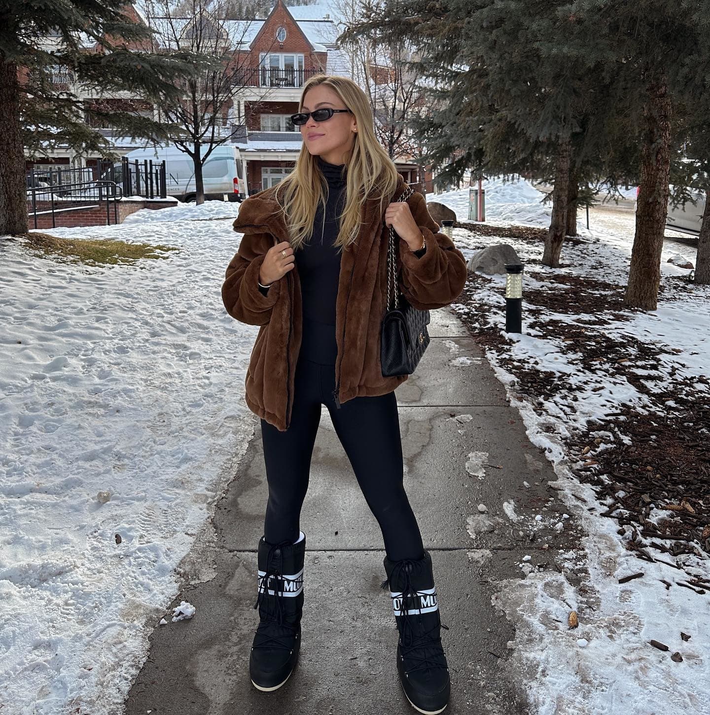 @marillewellyn wearing a mid-length faux fur jacket with a pair of warming black leggings and chunky snow boots while posing in a sidewalk lined with snow and pine trees.  