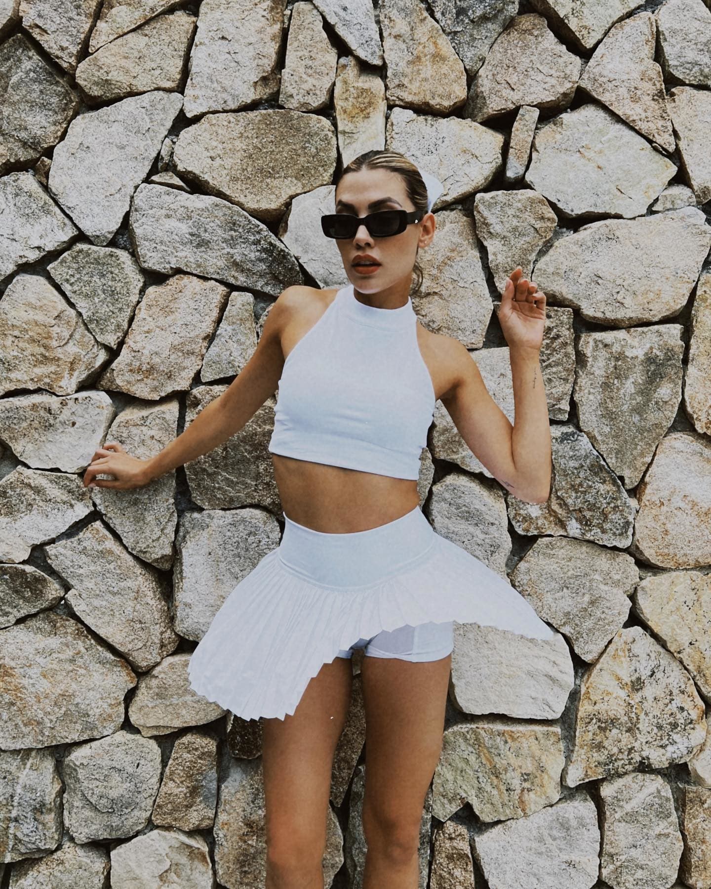 @michellesalasb wearing a white high neck cropped bra tank with a white pleated tennis skirt while posing in front of a rock wall.  