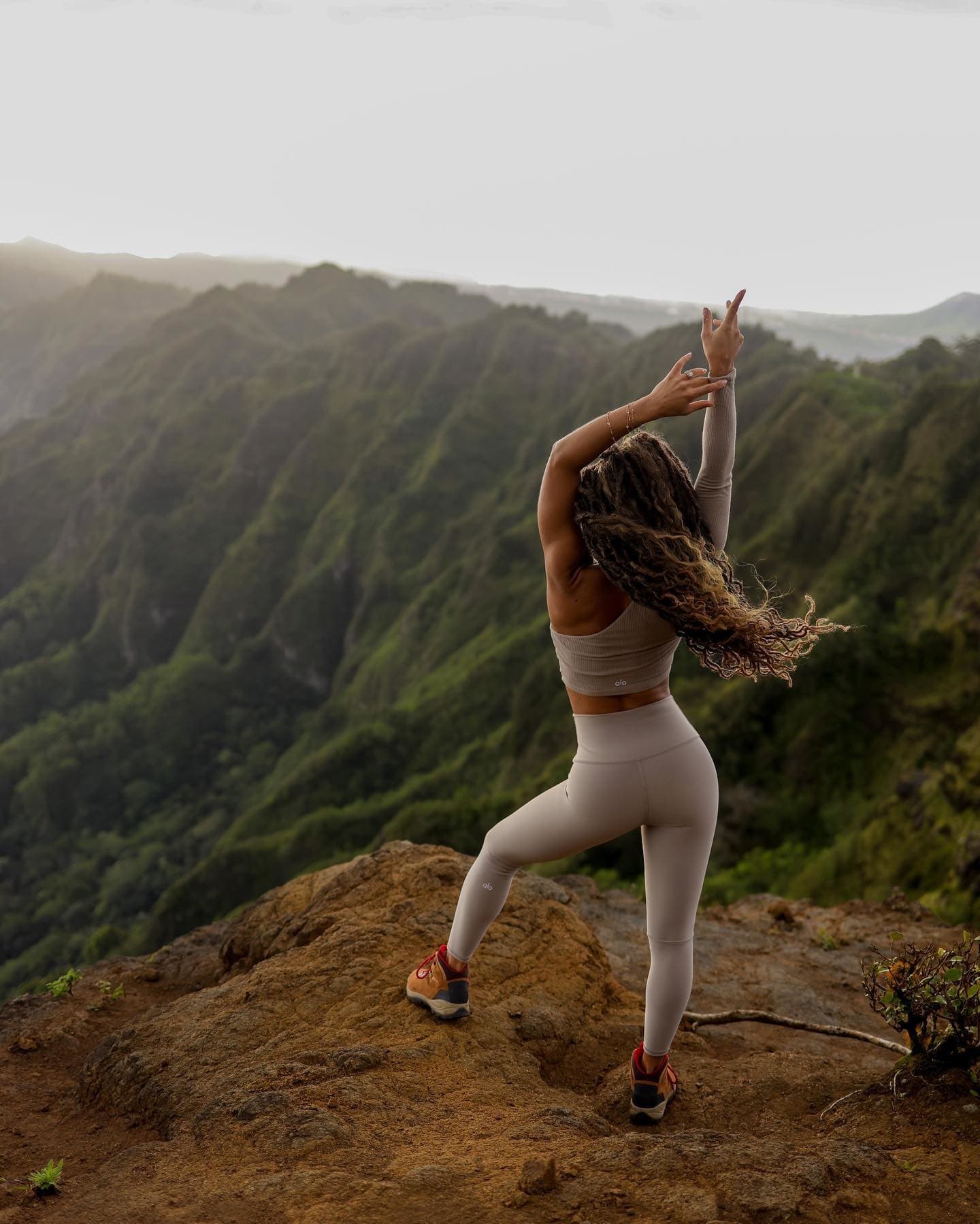 @phyllicia.bonanno wearing a pair of high-waisted leggings with a matching one-sleeved long sleeve shirt while posing at the top of a mountain at sunrise  