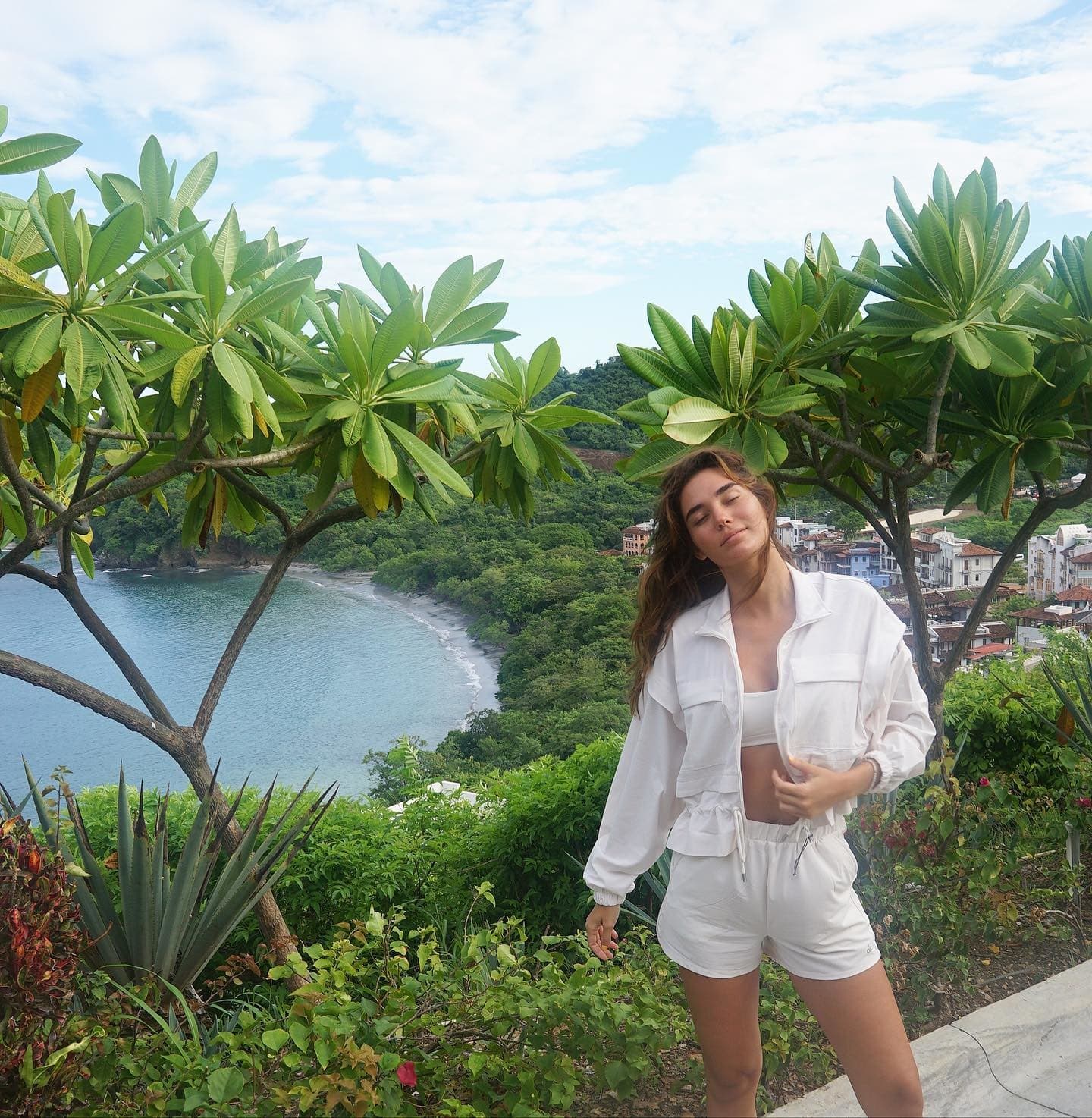 @rachellvallori wearing a pair of white crinkle fabric shorts with a matching cropped jacket and sports bra while posing on a hilltop overlooking a beach and neighboring town.  