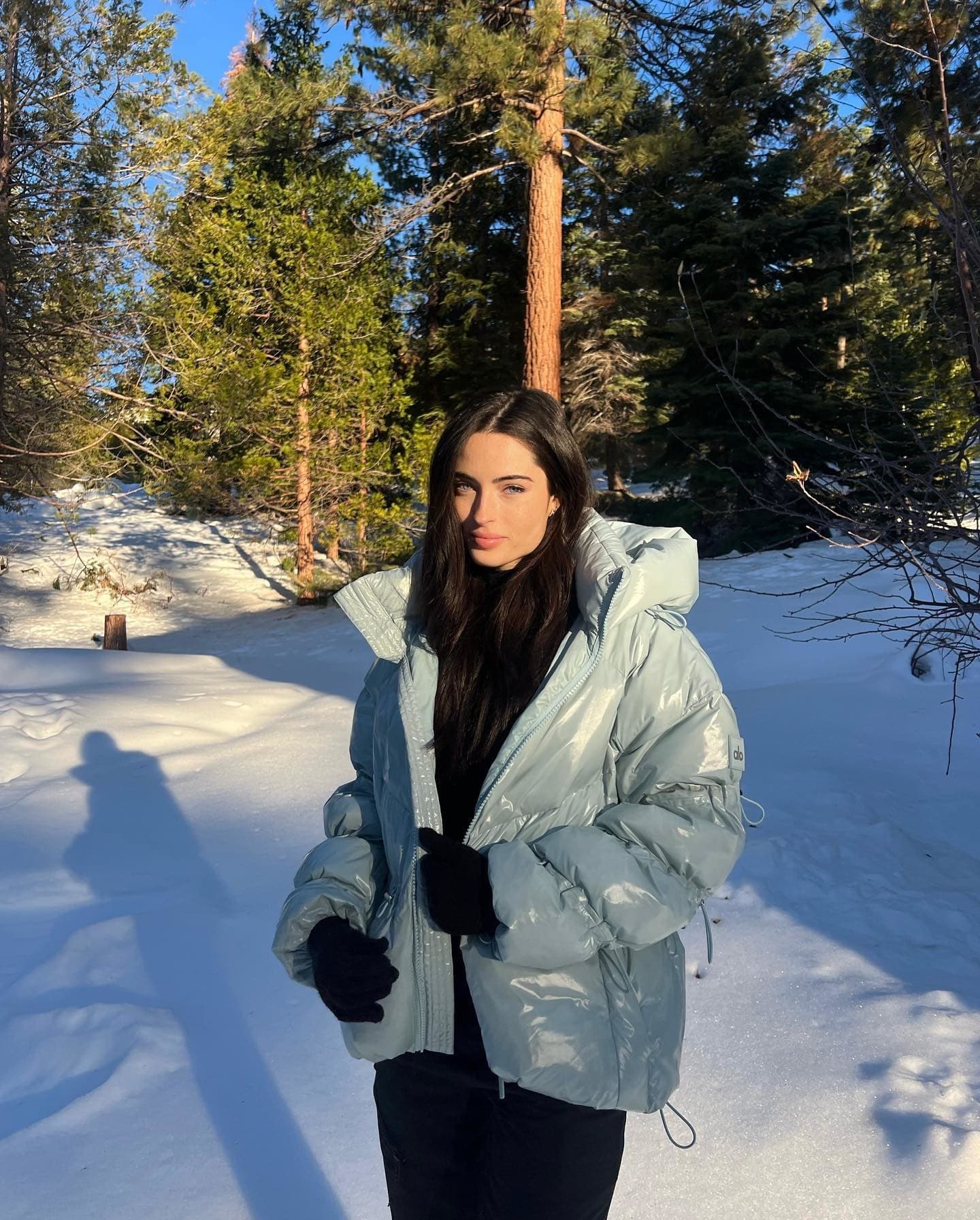 @reneemurden wearing a reflective puffer jacket in light blue with a micro sherpa sweat set underneath standing in fresh snow with pine trees in the background.  