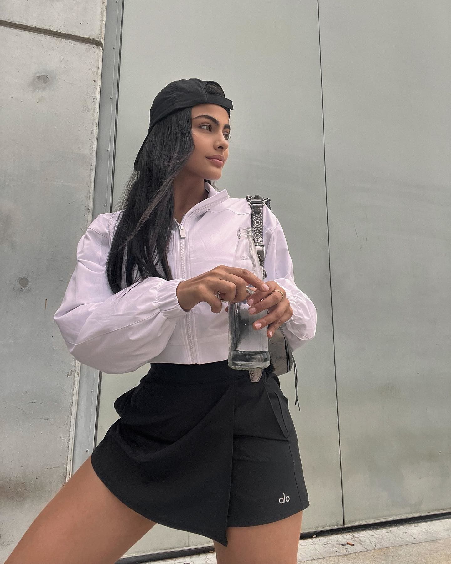@saraorrego wear a black high-waisted activewear skort with a cropped full-zip jacket while posing with a glass water bottle against a concrete wall. 
