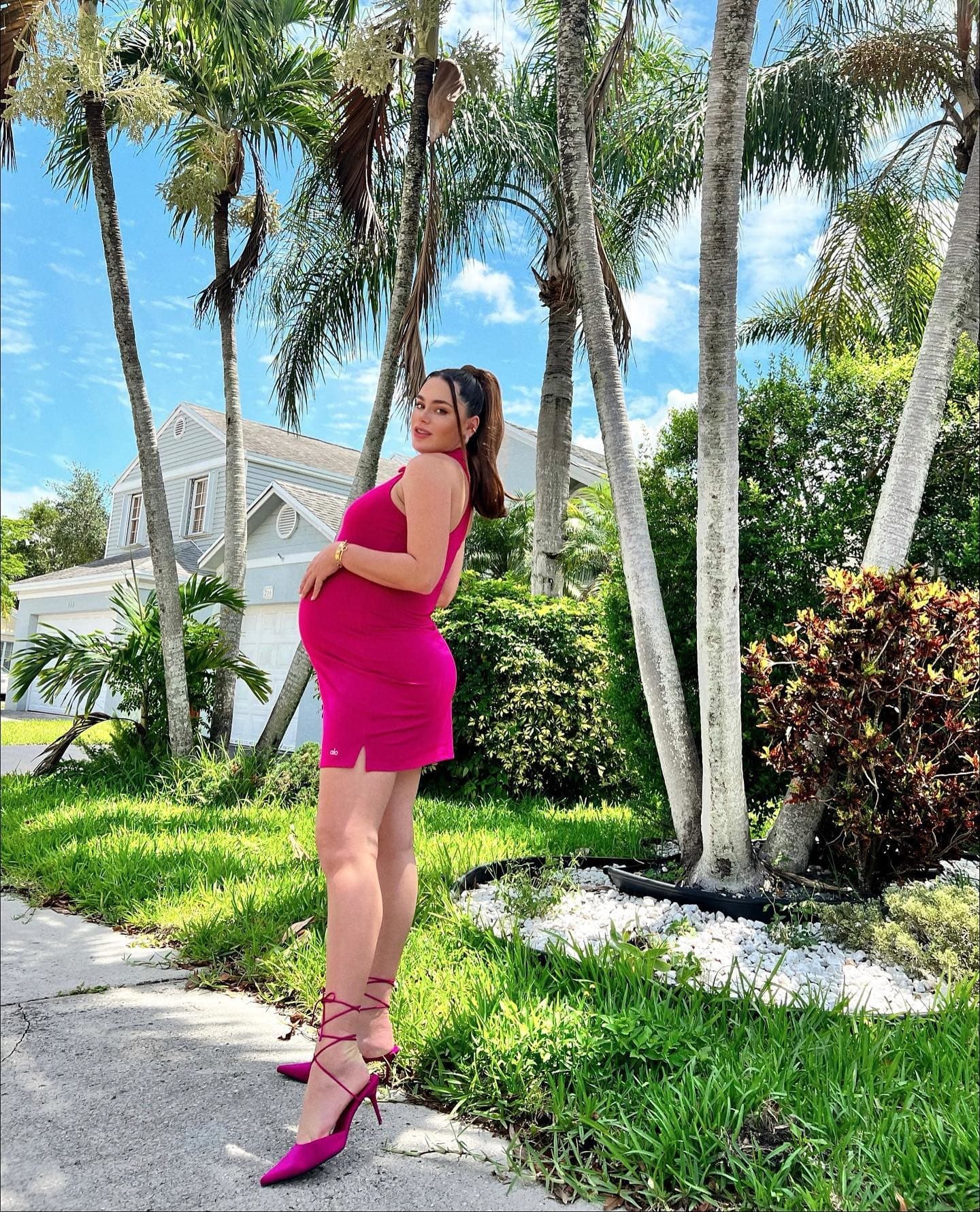 @stephistegman wearing a magenta collared tennis dress with a pair of matching high heels while posing with her hands on her pregnant belly in front of a patch of palm trees.  