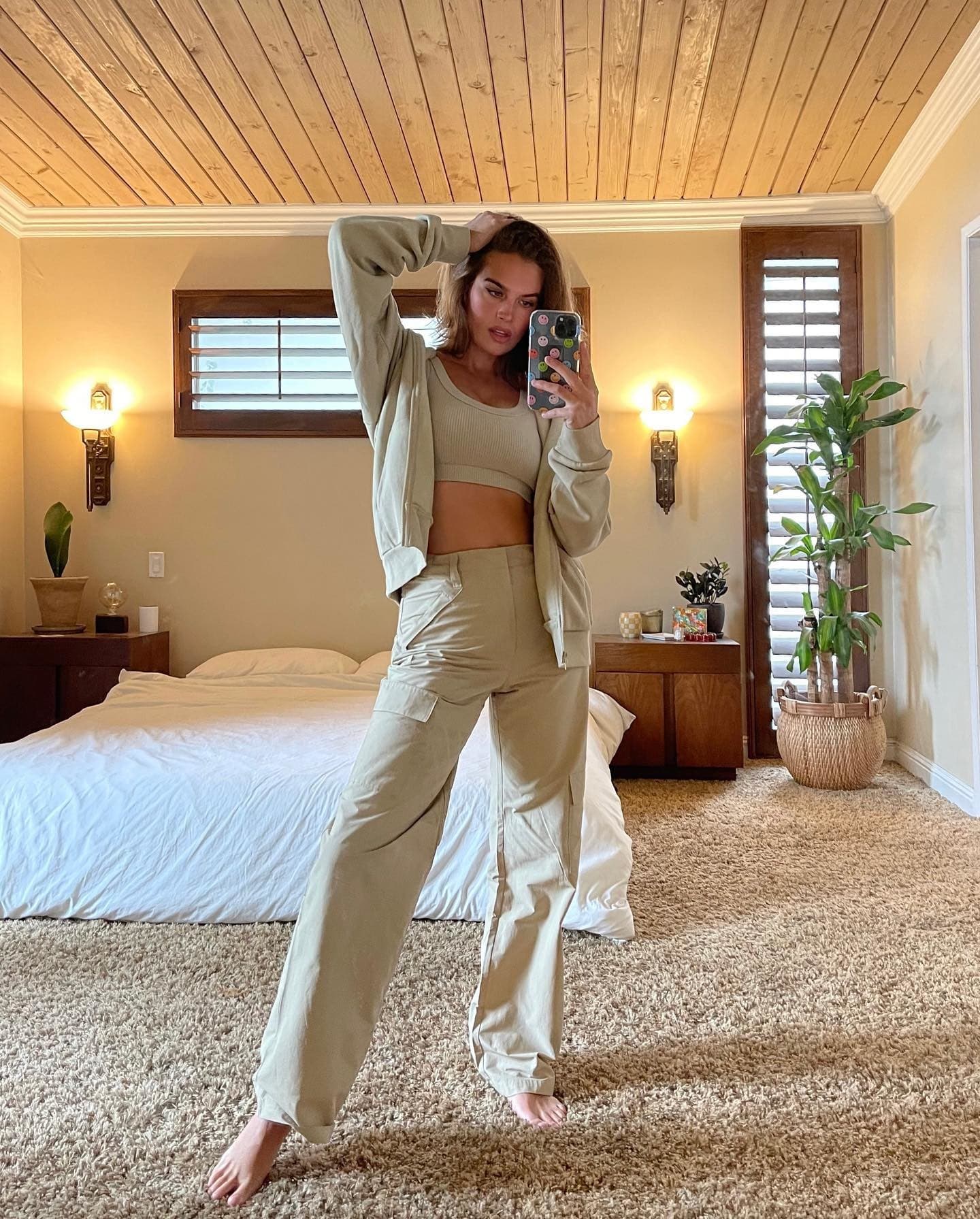 @stormibree wearing a pair of tan trousers with a sports bra and oversized zip-up hoodie while posing for a mirror picture within a bedroom. 